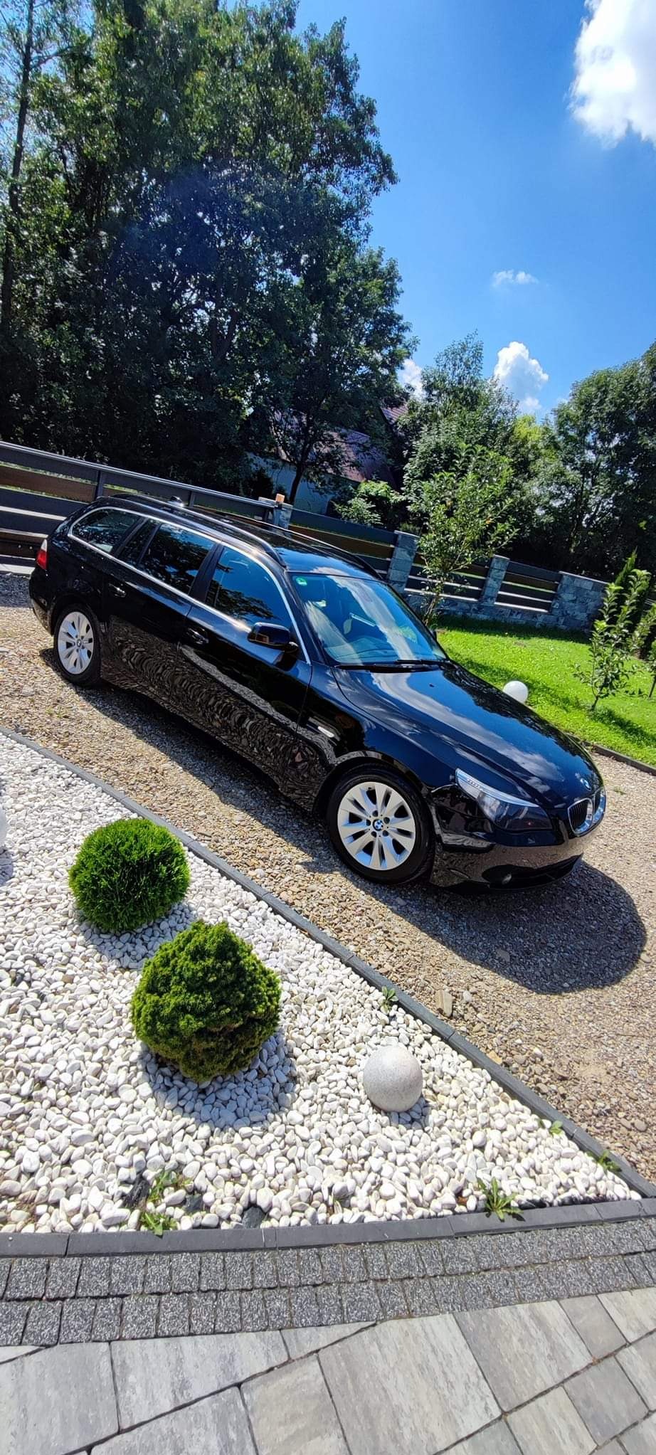 Czarny samochód kombi zaparkowany na białym żwirze, z dwoma kulistymi krzewami i kamienną kulą dekoracyjną w tle, widok na zadbany ogród i błękitne niebo.