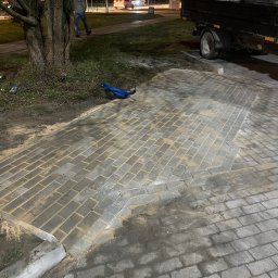 Vova Bruk - Świeżo ułożona szara kostka brukowa z widocznymi spoinami wypełnionymi piaskiem, obok krawężnika i drzewa, w tle przyczepa samochodowa i przechodzień w nocnej scenerii miejskiej.