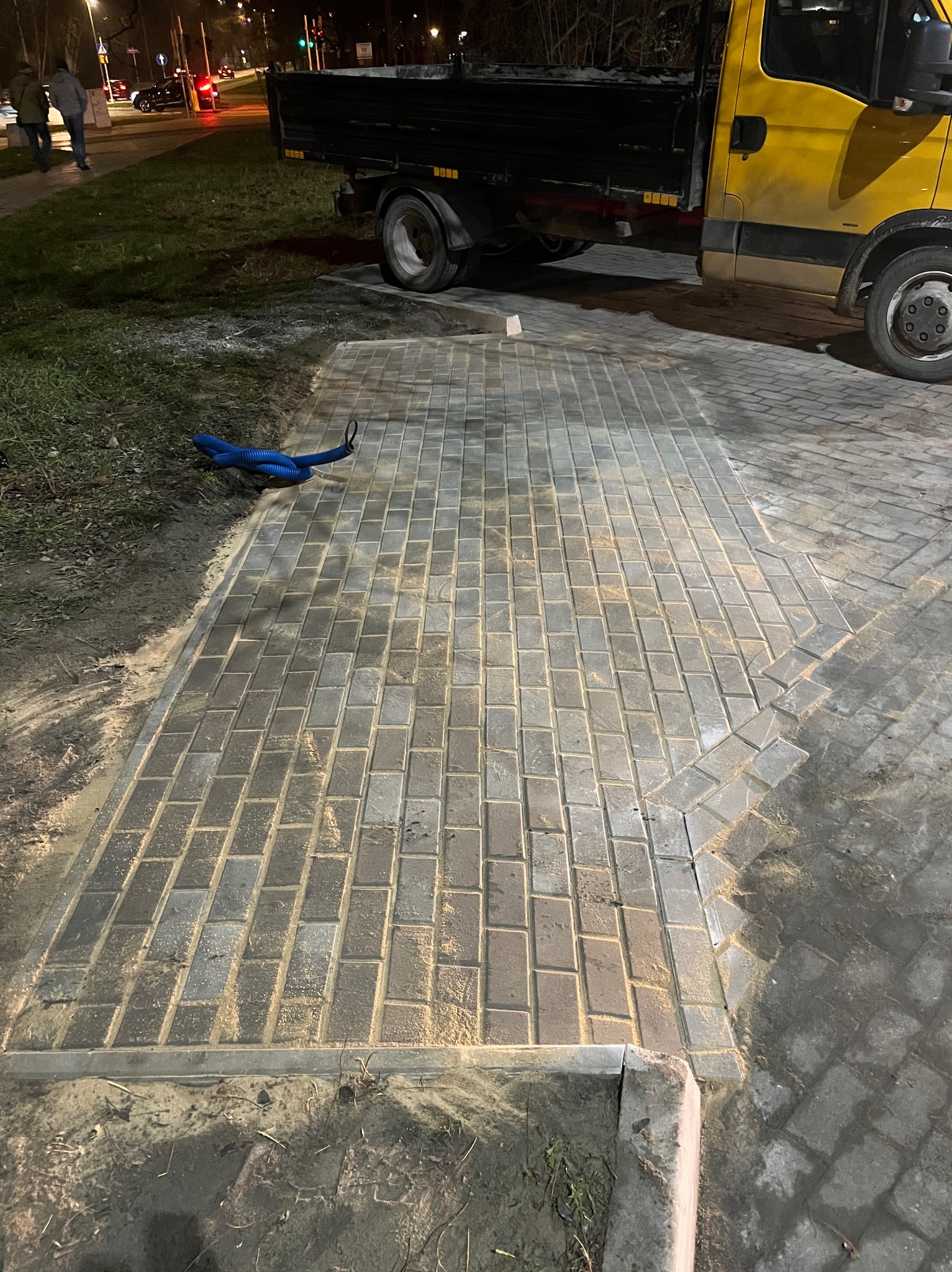 Fragment nowo ułożonej, prostokątnej nawierzchni z kostki brukowej, z widocznym żółtym samochodem ciężarowym w tle i niebieskim przewodem na boku, częściowo zasypana piaskiem.