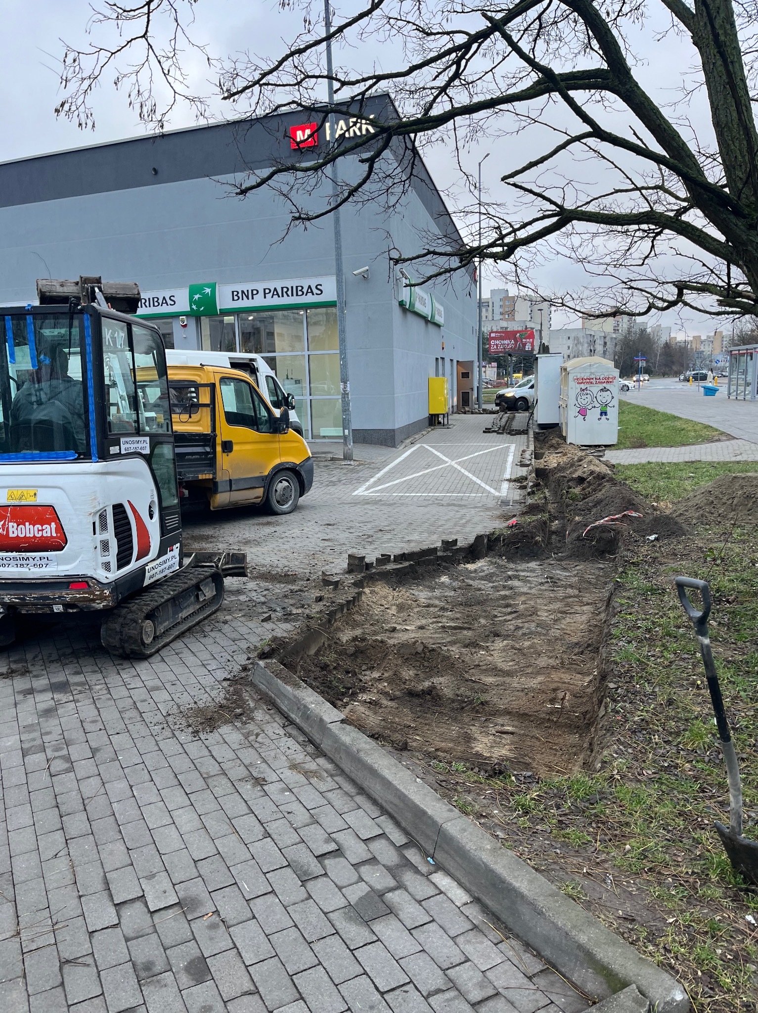 Mini koparka Bobcat i żółty samochód dostawczy na tle budynku z bankiem BNP Paribas, obok wykopany rów w ziemi, przygotowanie podłoża pod chodnik z kostki brukowej.