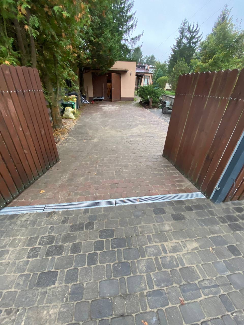 Ułożona kostka brukowa na podjeździe z widocznym odpływem liniowym, otwierana drewniana brama w tle.