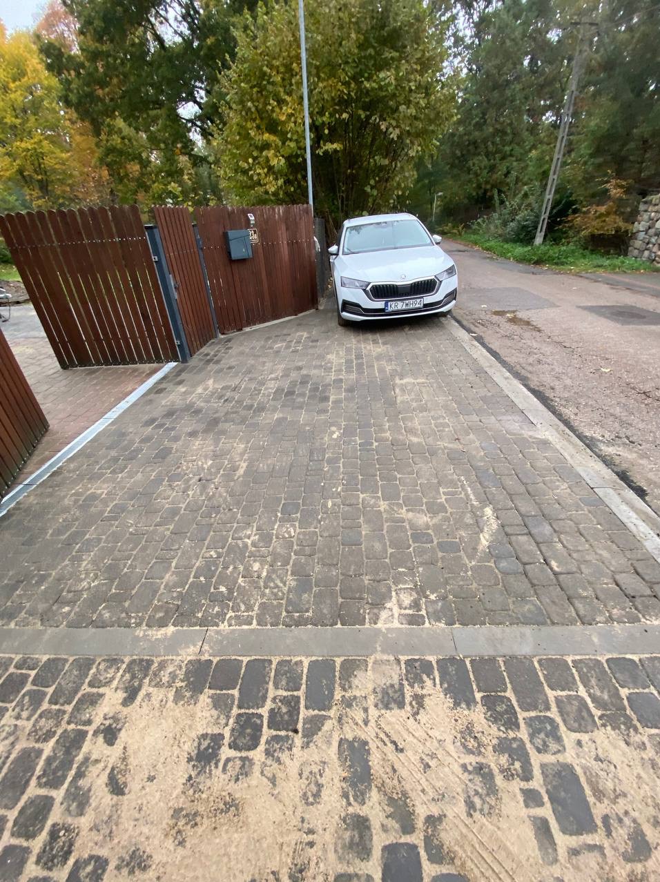 Ułożona kostka brukowa na podjeździe przed domem, widoczny biały samochód osobowy i drewniane ogrodzenie z bramą.