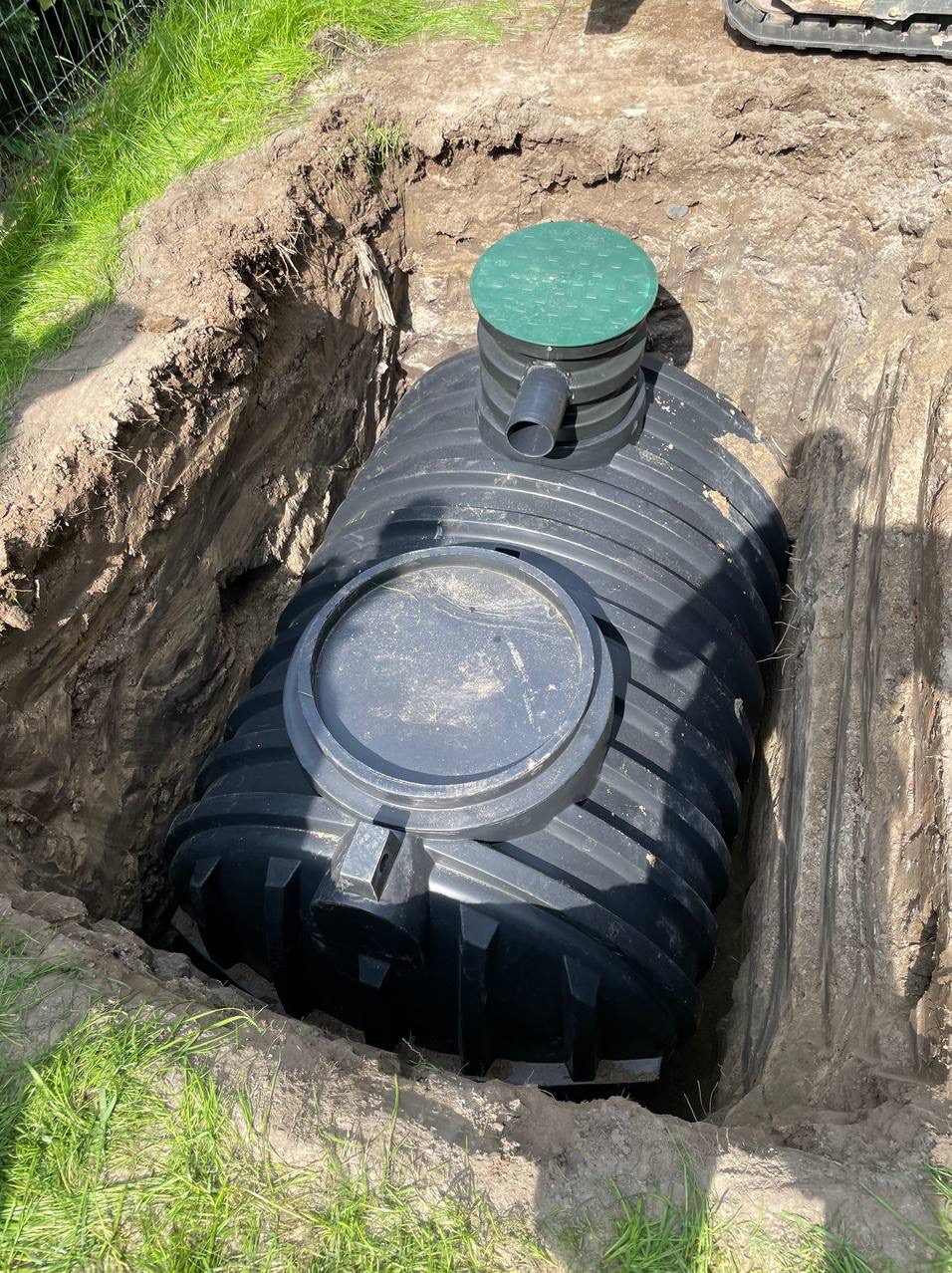 Czarny zbiornik na szambo z zieloną pokrywą częściowo zagłębiony w wykopie ziemnym, widoczne ślady po szpadlu na ścianach wykopu.