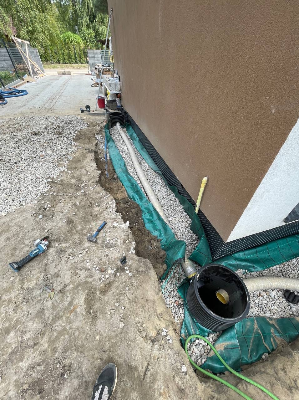 Rów przy fundamencie budynku z ułożoną rurą drenażową, obsypaną żwirem i zabezpieczoną geowłókniną; widoczny czarny zbiornik drenażowy, narzędzia budowlane oraz fragment buta osoby fotografującej.