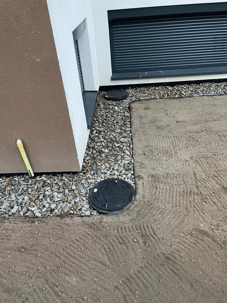 Przygotowany teren pod przyszły trawnik z widocznymi studzienkami kanalizacyjnymi i obsypką z kamieni ozdobnych przy ścianie budynku.