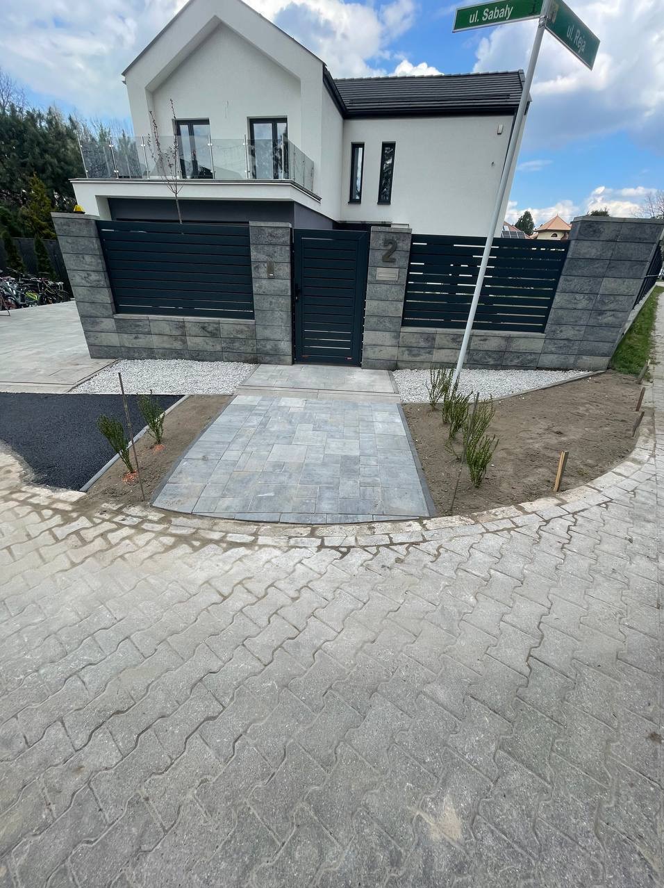 Nowa, szara kostka brukowa ułożona na podjeździe do nowoczesnego domu z ciemnozielonym ogrodzeniem, widoczny fragment asfaltowej drogi i znak z nazwami ulic.