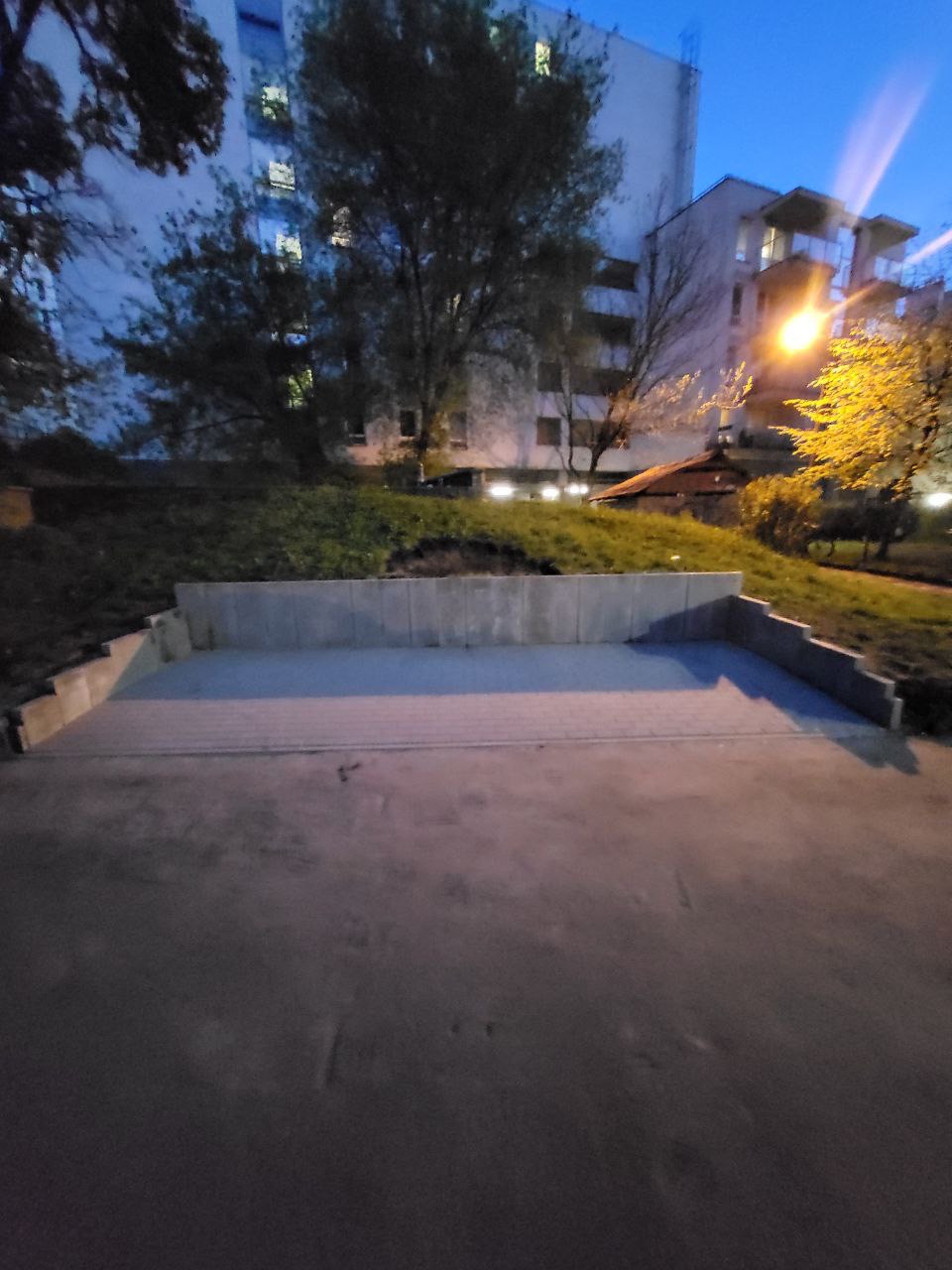 Nowo ułożona, szara kostka brukowa tworząca plac przed budynkiem mieszkalnym, otoczona niskim, betonowym murkiem oporowym. Zdjęcie wykonane o zmierzchu.