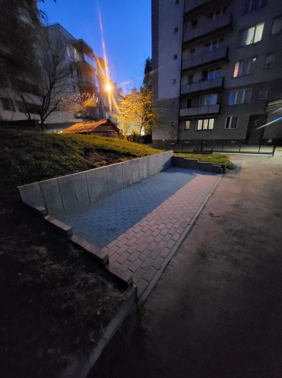 Ukończony wjazd do garażu podziemnego wyłożony szarą kostką brukową, otoczony betonowym murem oporowym, sfotografowany o zmierzchu na tle budynków mieszkalnych i oświetlenia ulicznego.