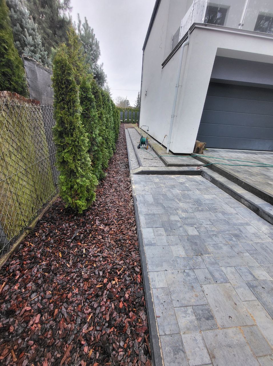 Ułożona szara kostka brukowa prowadząca do nowoczesnego domu z garażem; obok rabata z korą i rząd tuj wzdłuż ogrodzenia.