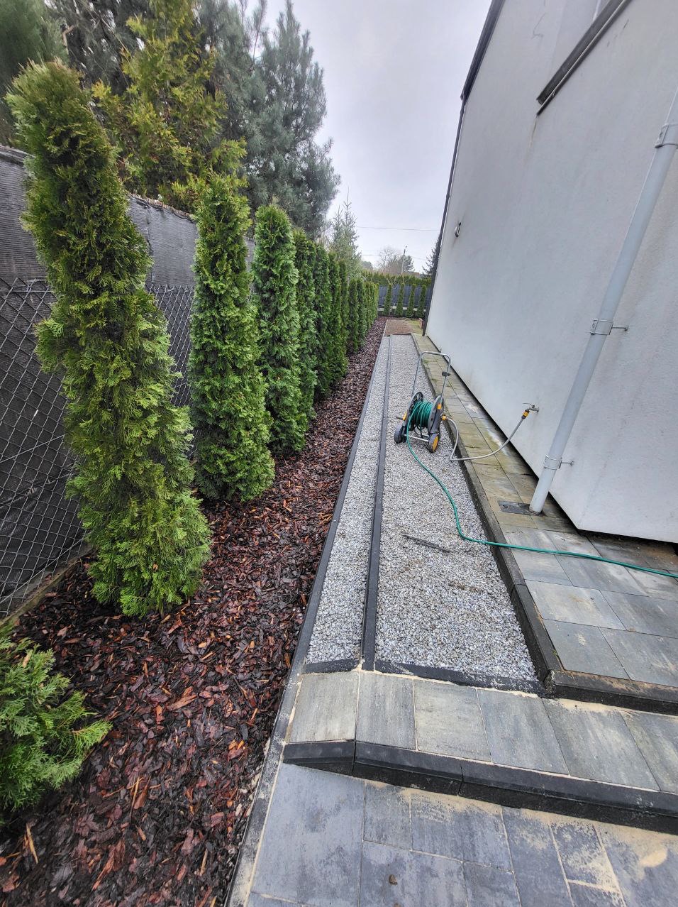 Wykończony fragment ogrodu z równo ułożonymi szarymi płytami chodnikowymi, wąskim pasem drobnego żwiru oddzielonym czarnym obrzeżem i rzędem posadzonych tuj wzdłuż ogrodzenia z siatki.