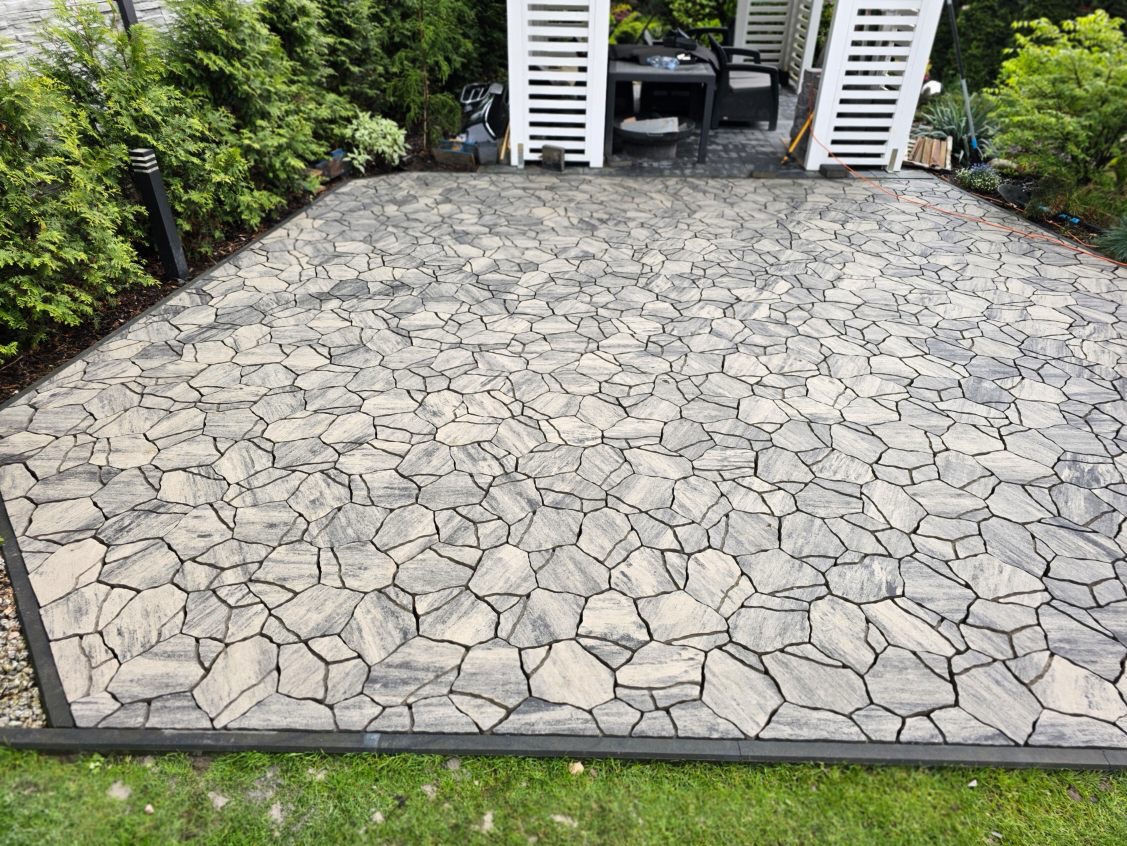 Wykończony taras z nieregularnej, szarej kostki brukowej, obrzeża z ciemnego tworzywa, zieleń w tle i biała altana. Widok z góry.