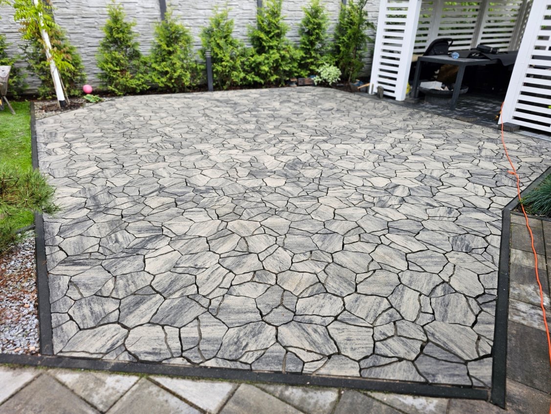 Ukończona nawierzchnia z nieregularnej, szarej kostki, obramowana czarnym brzegiem. W tle zielone krzewy i biała altana. Widok na ogród z bliska.