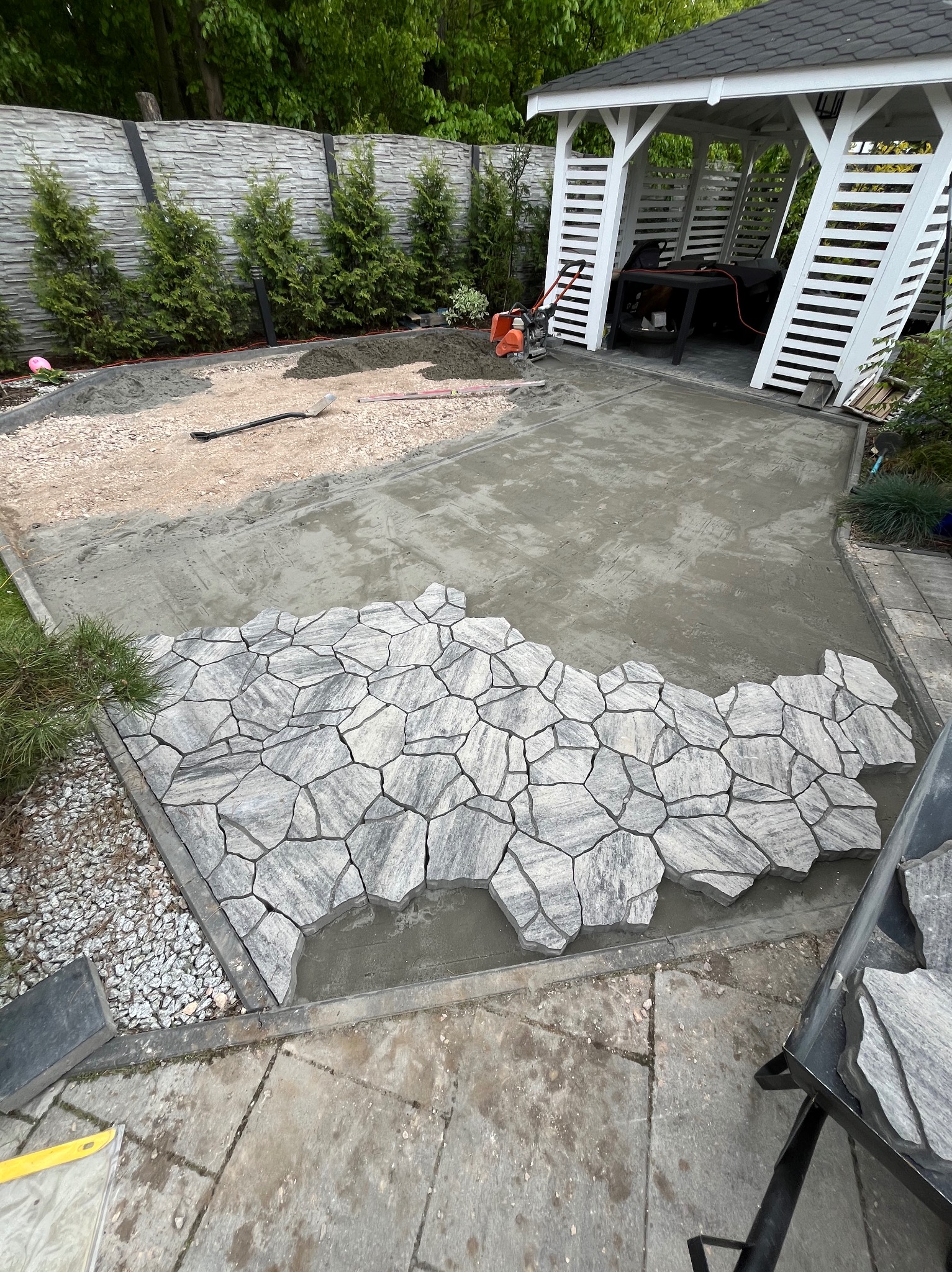 Fragment układania szarej kostki o nieregularnym kształcie na betonowej wylewce, w tle widoczny biały altanka i przygotowany teren pod dalsze prace brukarskie.