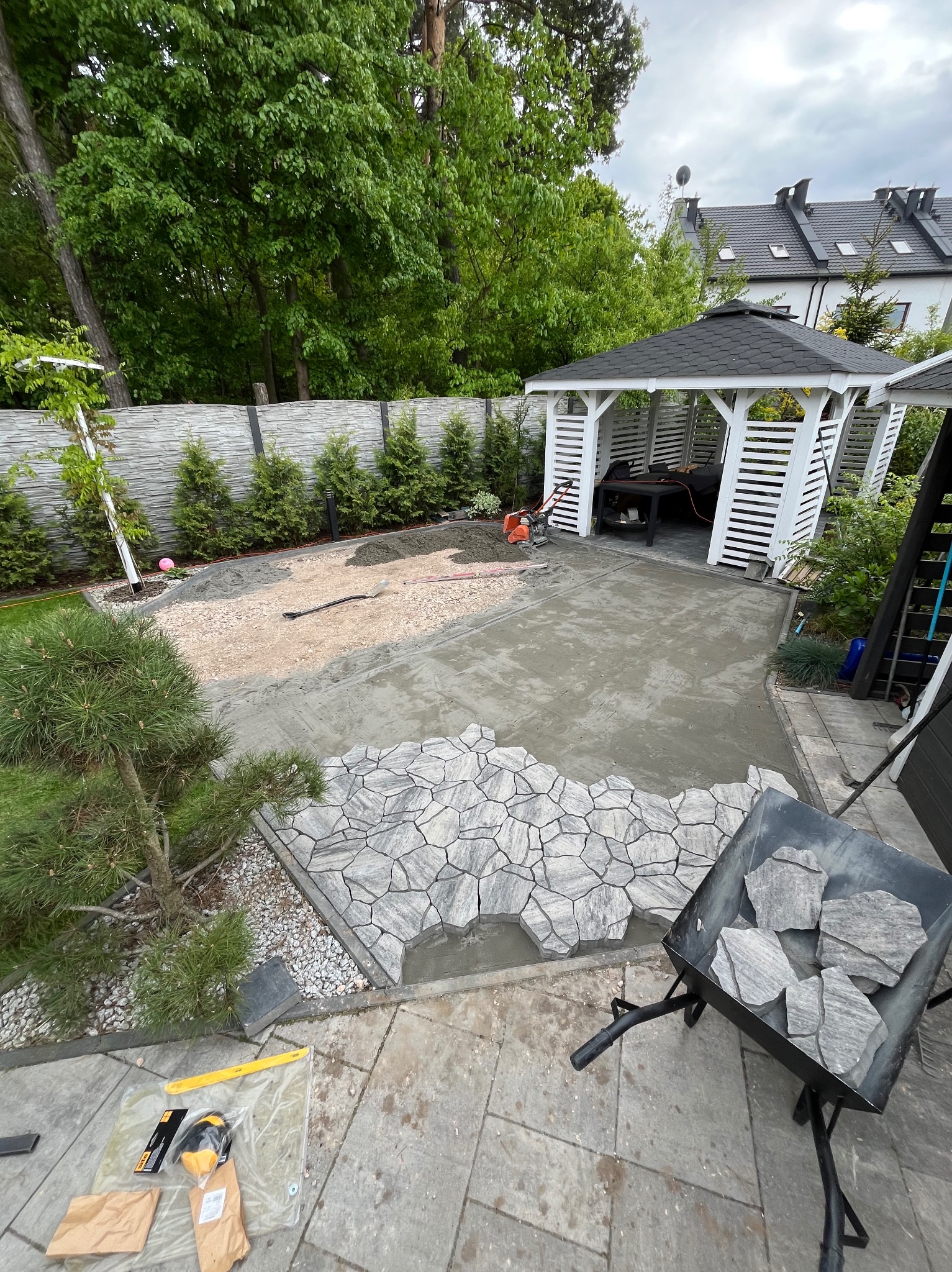 Układanie nieregularnej kostki brukowej na zaprawie w ogrodzie z altaną. Widoczny wózek z kostką, narzędzia i przygotowany teren pod dalsze prace.