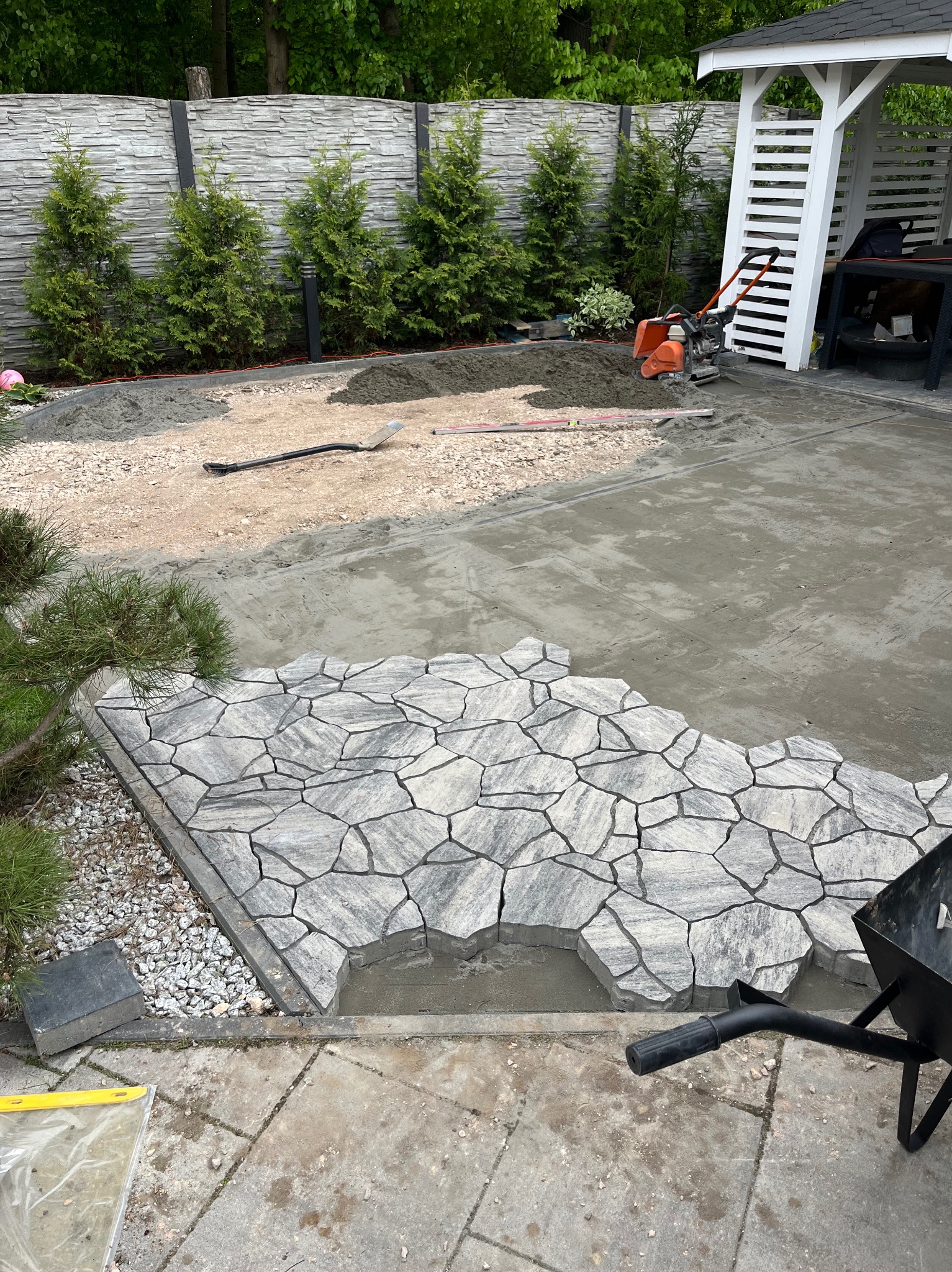 Fragment układania nieregularnej kostki brukowej w ogrodzie. Widoczny etap prac: przygotowane podłoże, częściowo ułożona kostka, narzędzia i taczka. Tło: ogrodzenie i roślinność.
