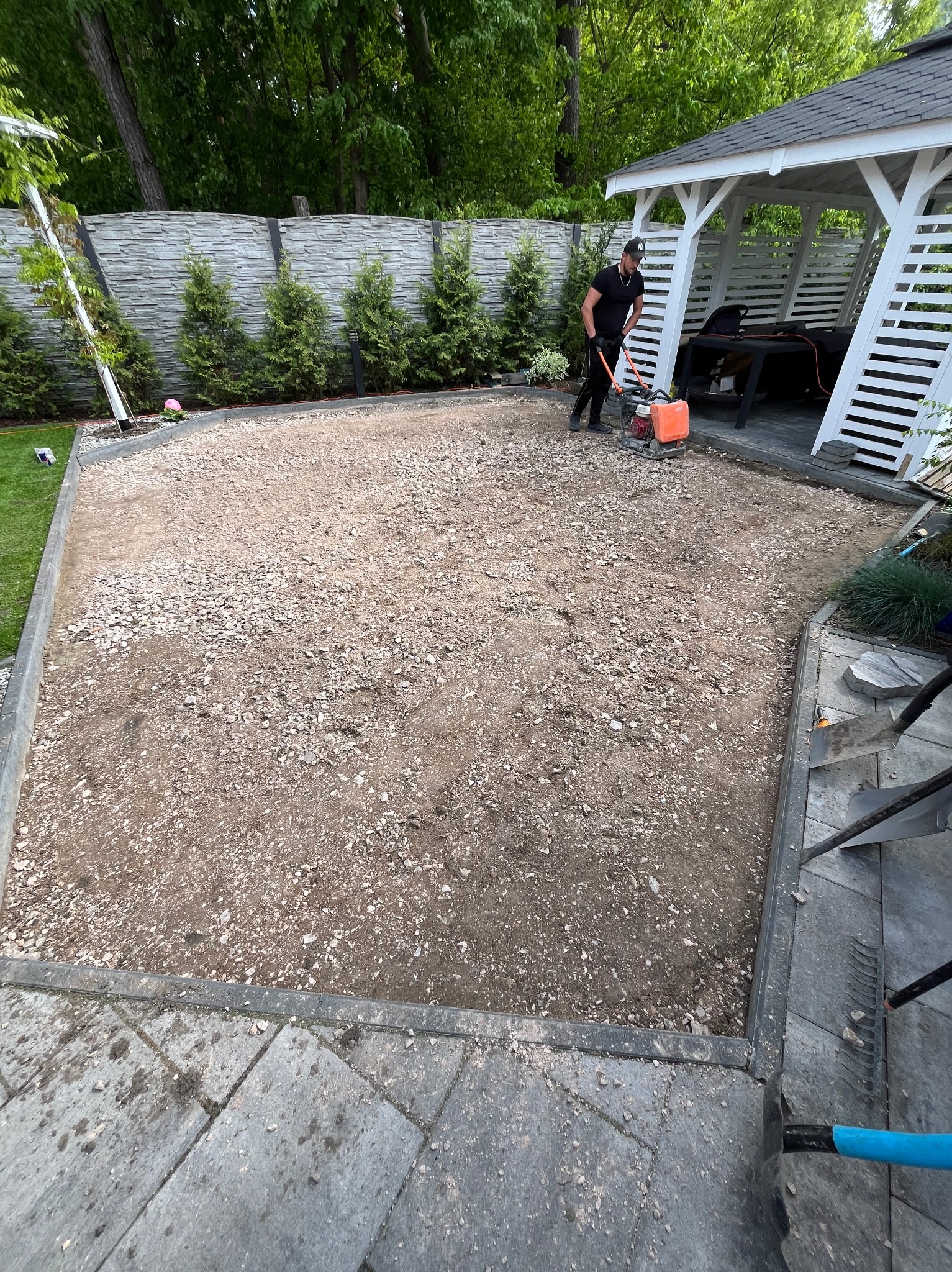 Prace przygotowawcze podłoża pod kostkę brukową: ubijanie ziemi zagęszczarką spalinową. Widoczne obrzeża, częściowo ułożona kostka, altana ogrodowa w tle.