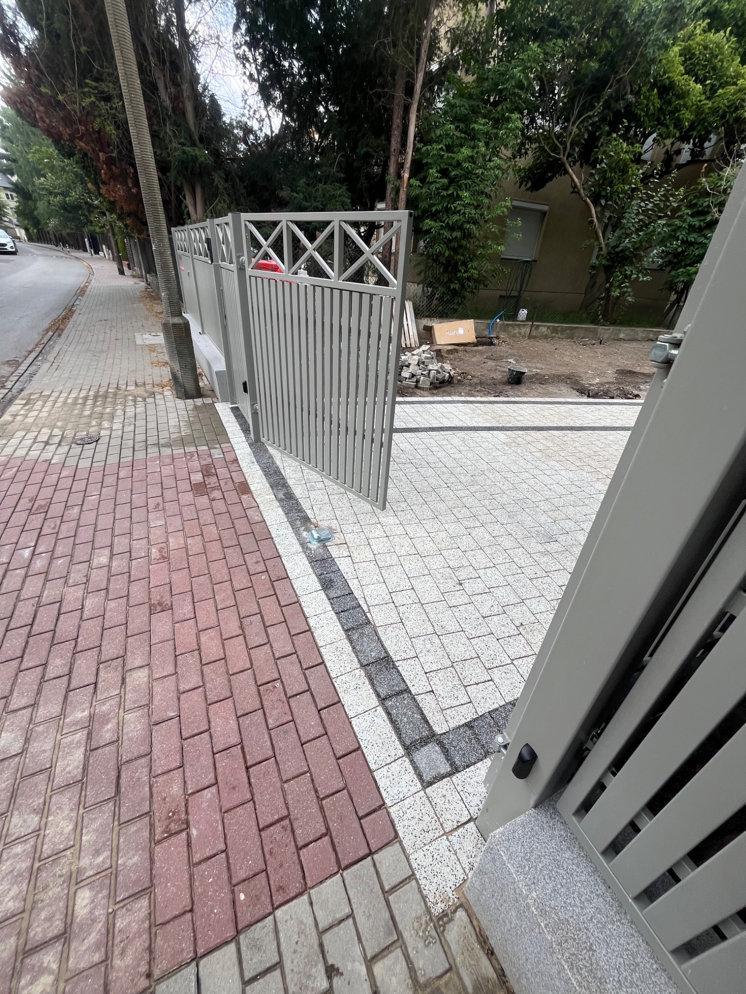 Nowa, jasna kostka brukowa ułożona na podjeździe z szarą bramą. Czerwona kostka chodnikowa po lewej. Trawa i drzewa w tle. Sezon wiosenno-letni.
