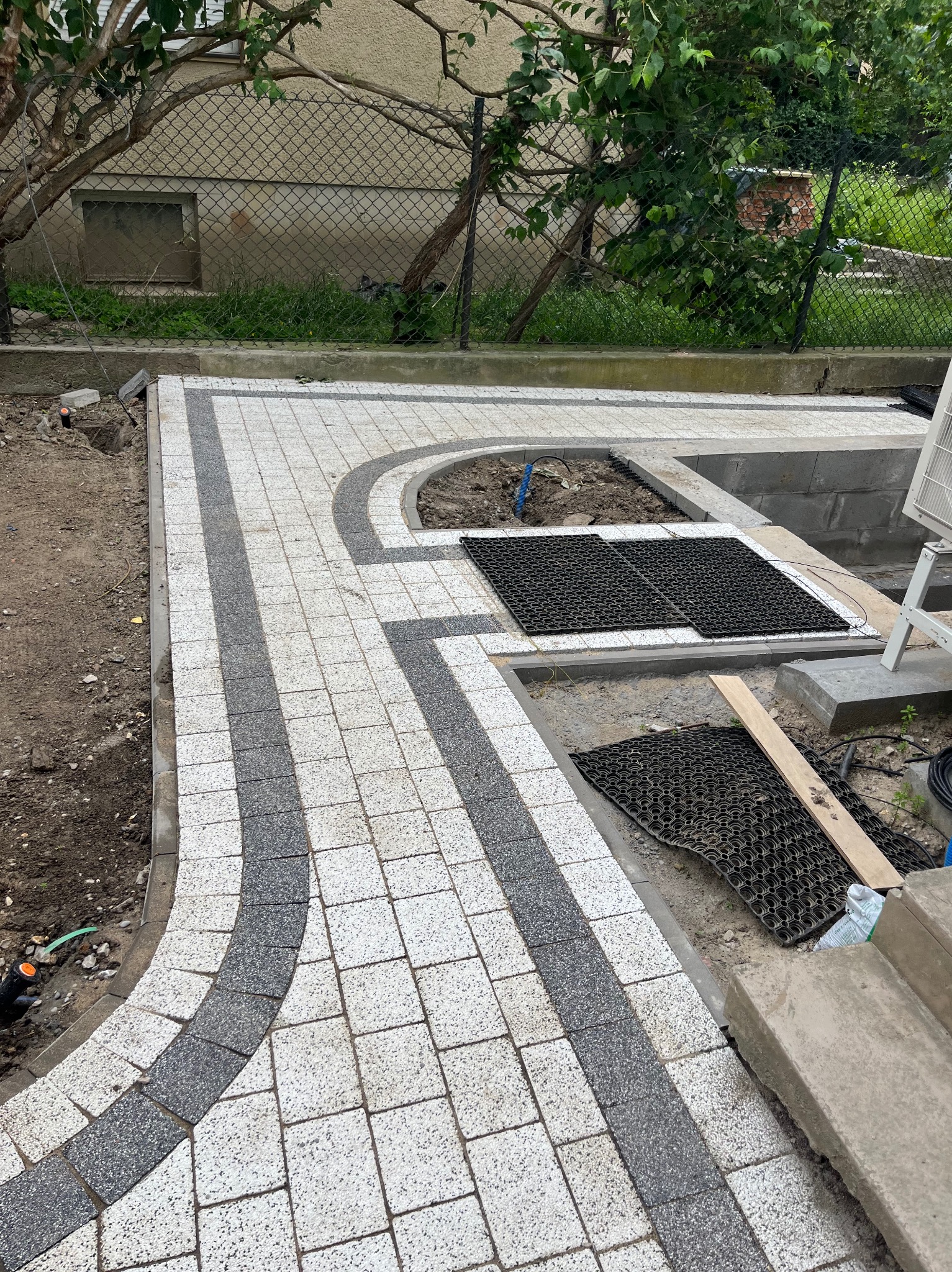 Ułożona kostka brukowa w szaro-białej kolorystyce, z widocznymi elementami przygotowania pod trawnik. Częściowo ukończona ścieżka w ogrodzie.