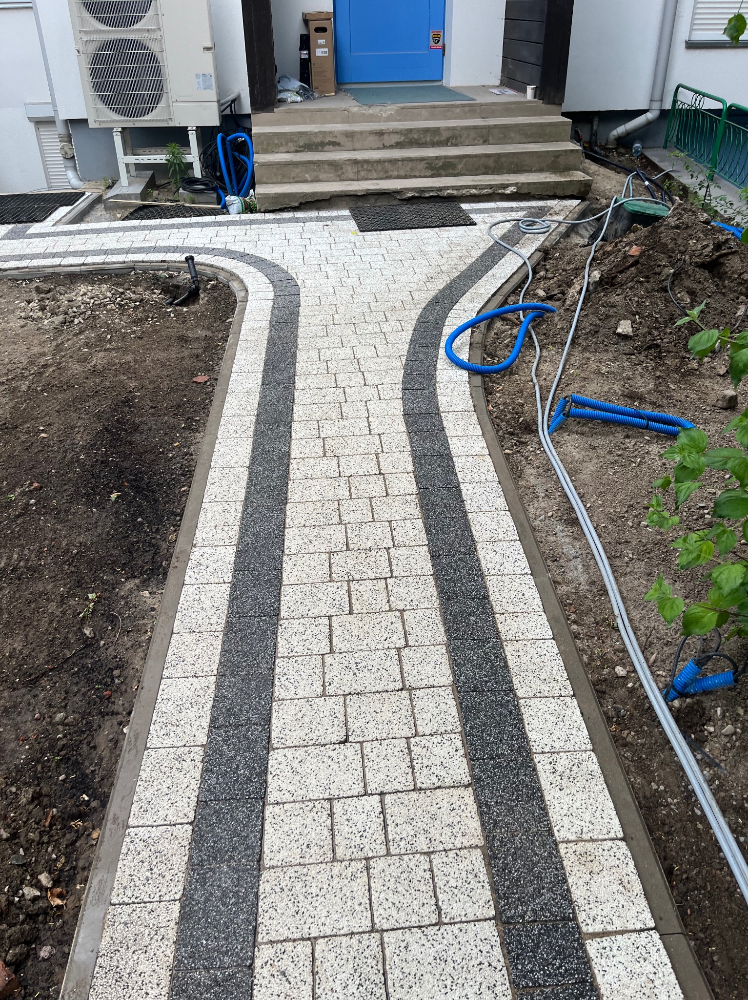 Ścieżka z kostki brukowej w dwóch odcieniach, rozwidlająca się w kierunku schodów wejściowych. Widoczne kable i rury wzdłuż krawędzi.