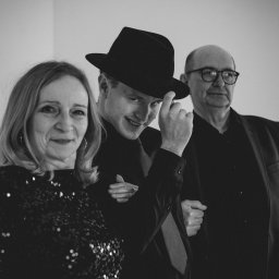 Fabryka Sztuki Mateusz Rudkowski - Czarno-białe zdjęcie przedstawia troje uśmiechniętych ludzi: kobietę w cekinowej bluzce, młodego mężczyznę w kapeluszu i starszego mężczyznę w okularach.
