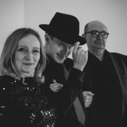 Czarno-białe zdjęcie przedstawia troje uśmiechniętych ludzi: kobietę w cekinowej bluzce, młodego mężczyznę w kapeluszu i starszego mężczyznę w okularach.