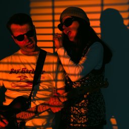 Fabryka Sztuki Mateusz Rudkowski - Duet muzyczny w okularach przeciwsłonecznych, mężczyzna gra na gitarze elektrycznej, kobieta śpiewa do mikrofonu, oświetleni pomarańczowym światłem z żaluzji, tworząc cienie na ścianie.