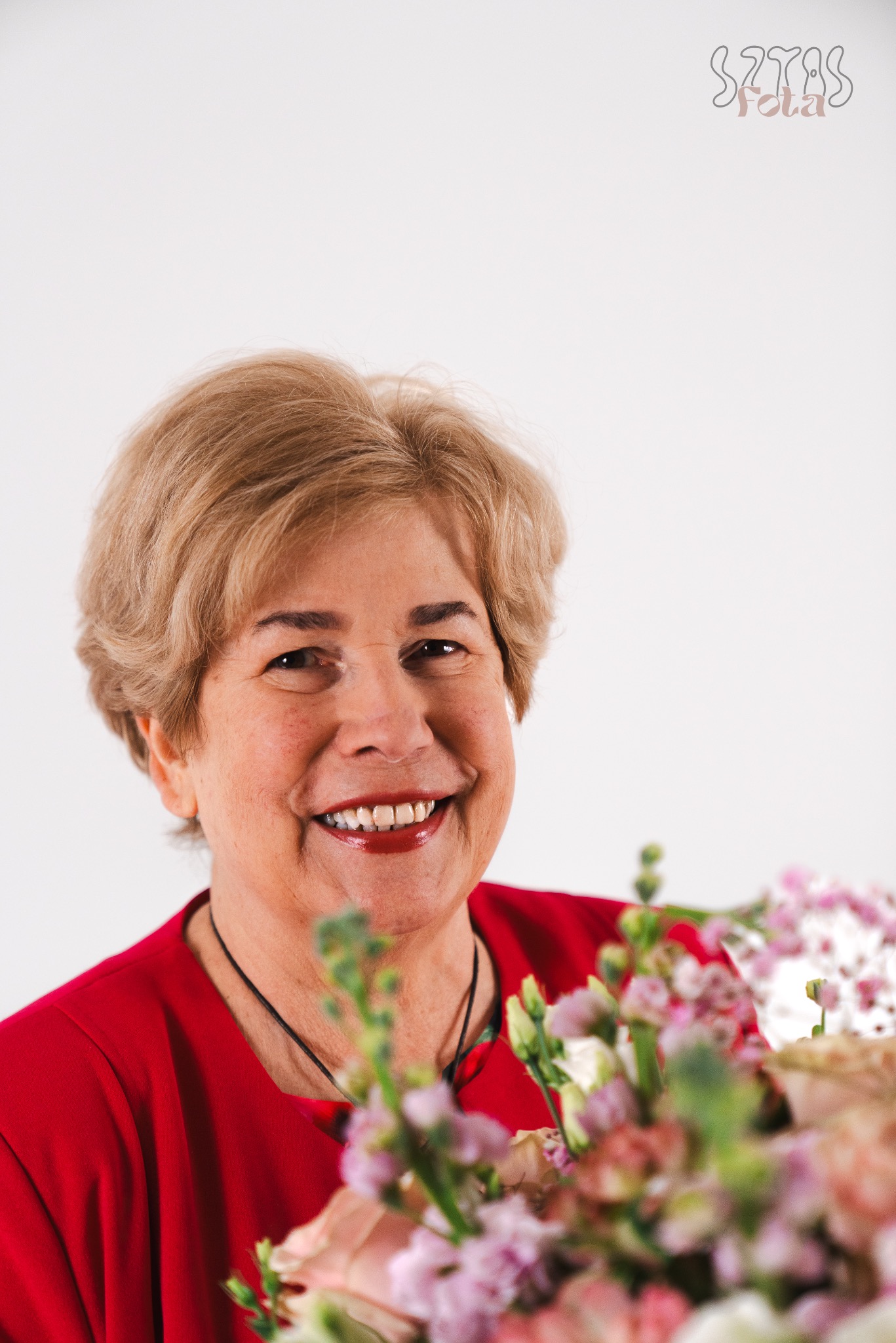 Portret uśmiechniętej kobiety w czerwonej bluzce, z bukietem kwiatów na pierwszym planie i logo fotografa w prawym górnym rogu.