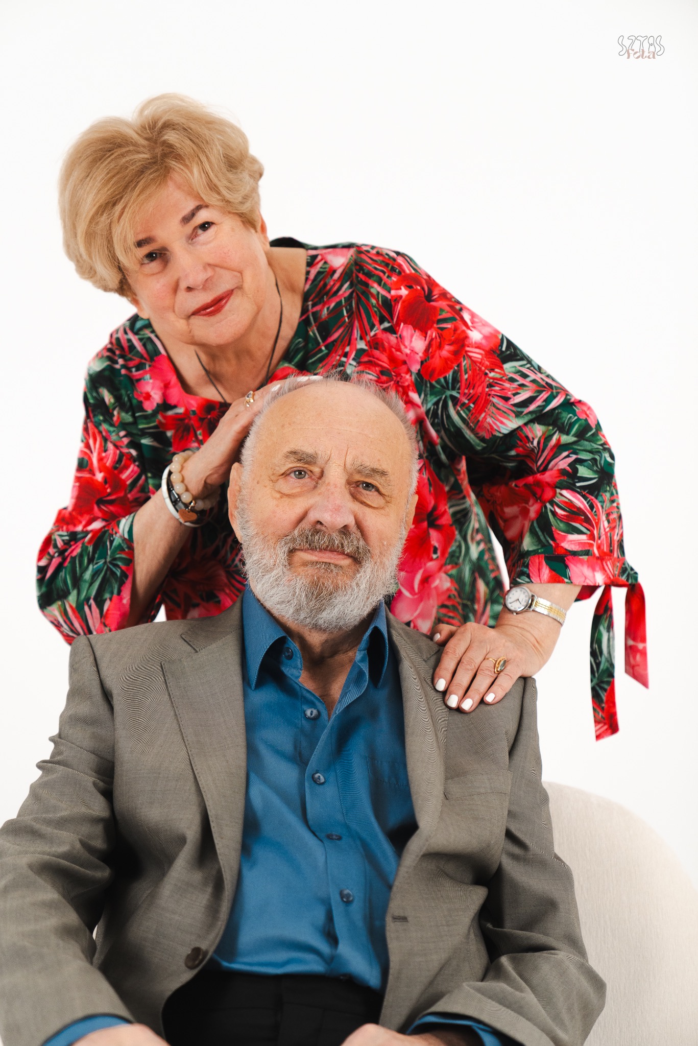 Portret eleganckiej, uśmiechniętej pary seniorów na jasnym tle: mężczyzna w szarej marynarce i niebieskiej koszuli siedzi, a kobieta w kwiecistej bluzce stoi za nim, opierając dłoń na jego głowie.