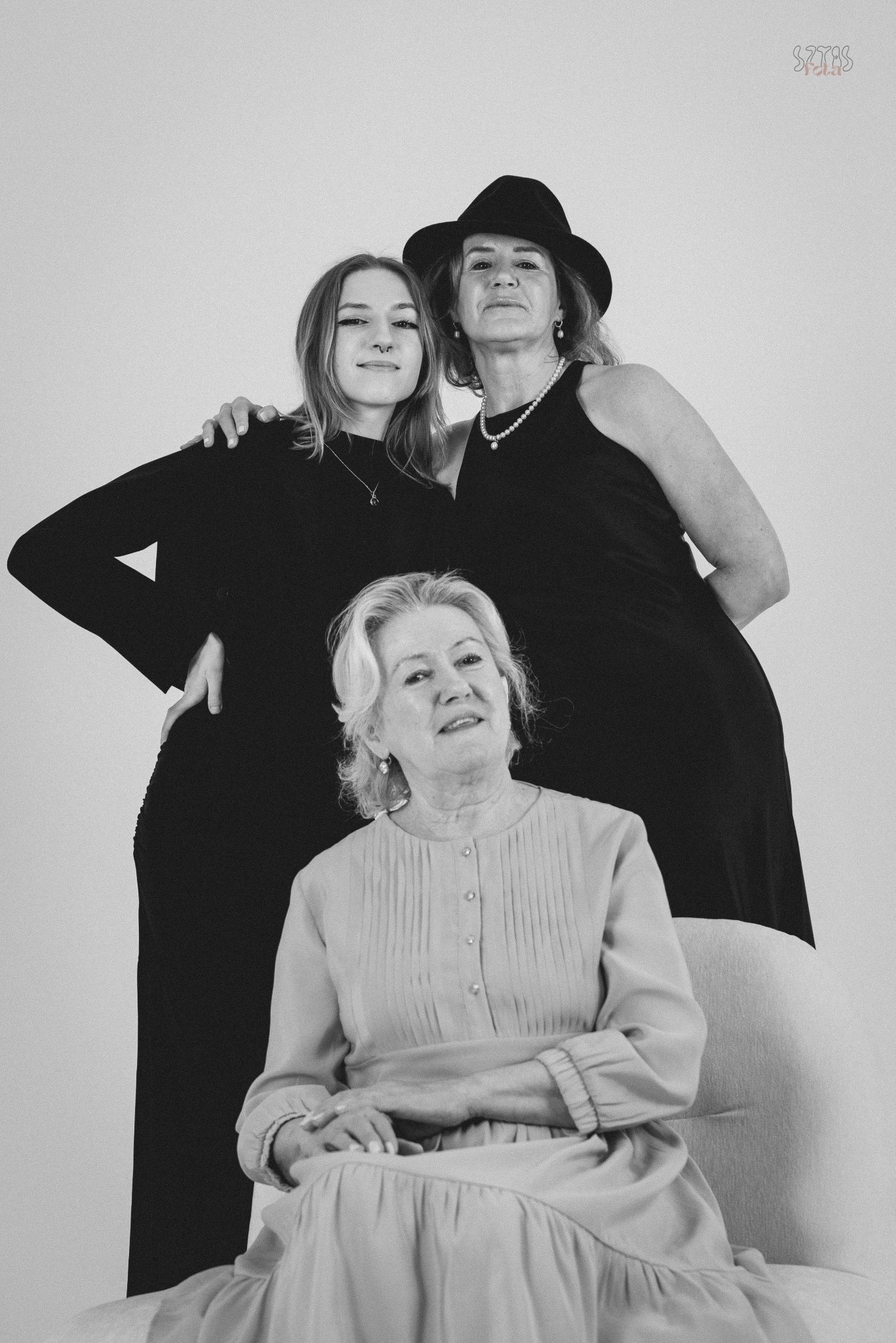 Czarno-białe portretowe ujęcie trzech kobiet w różnym wieku: siedząca starsza kobieta z dłońmi złożonymi na kolanach, za nią stojąca młodsza kobieta z piercingiem w nosie obejmująca ramieniem...