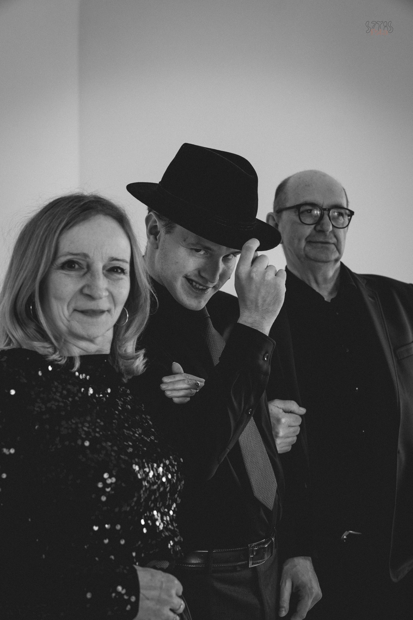Czarno-białe zdjęcie przedstawia troje uśmiechniętych ludzi: kobietę w cekinowej bluzce, młodego mężczyznę w kapeluszu i starszego mężczyznę w okularach.