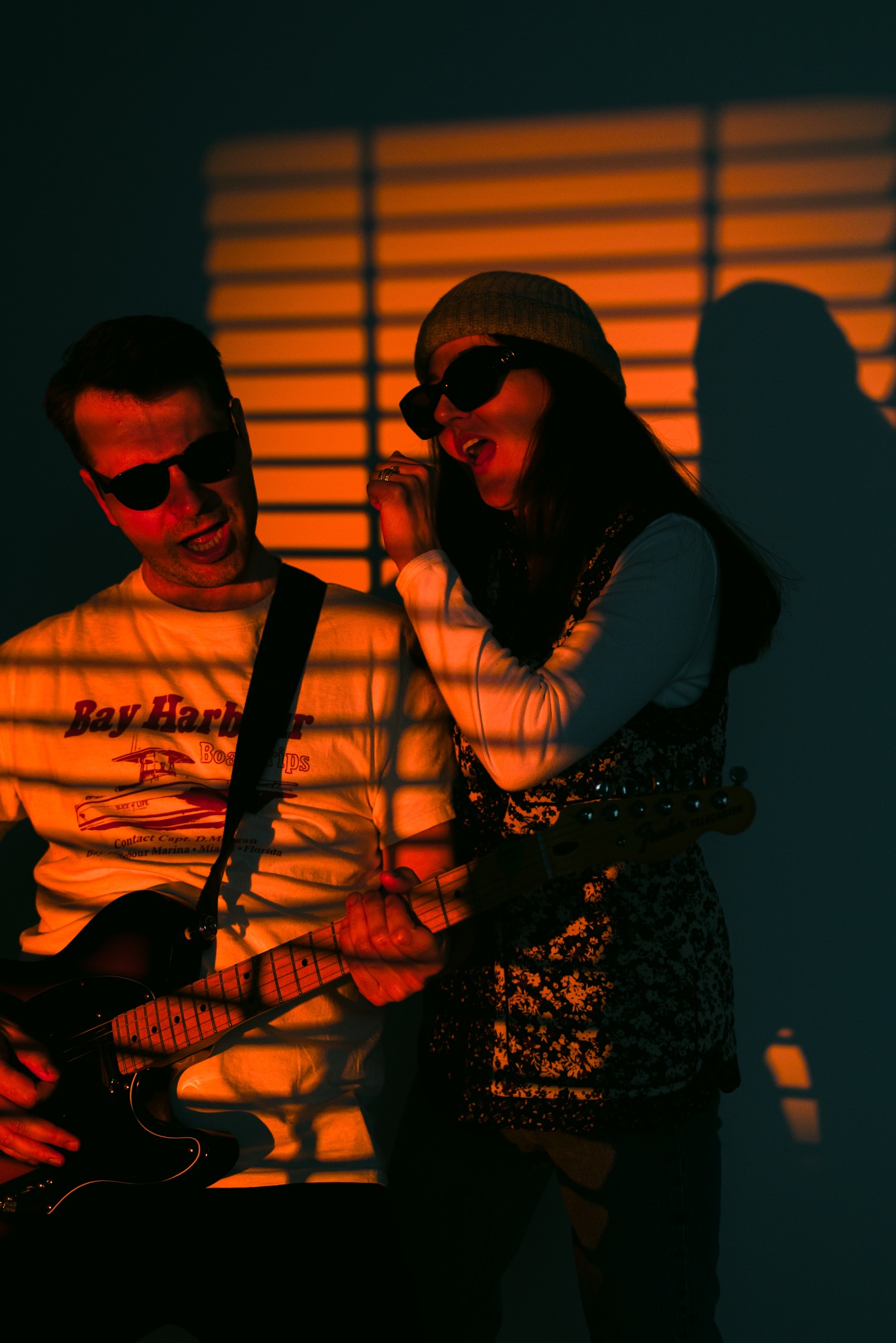 Duet muzyczny w okularach przeciwsłonecznych, mężczyzna gra na gitarze elektrycznej, kobieta śpiewa do mikrofonu, oświetleni pomarańczowym światłem z żaluzji, tworząc cienie na ścianie.