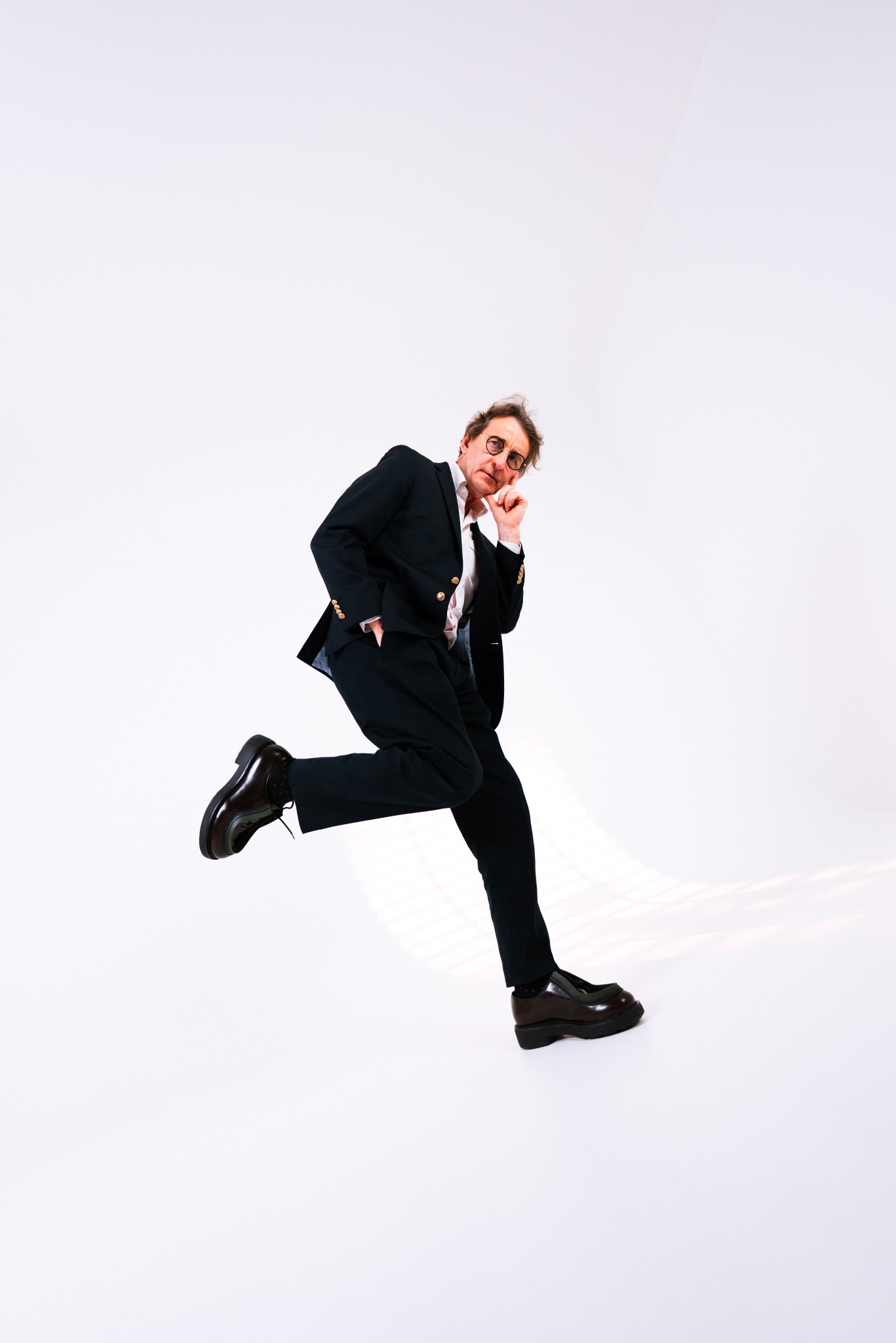 Mężczyzna w garniturze, w okularach, w dynamicznej pozie, z ręką pod brodą, na białym tle.