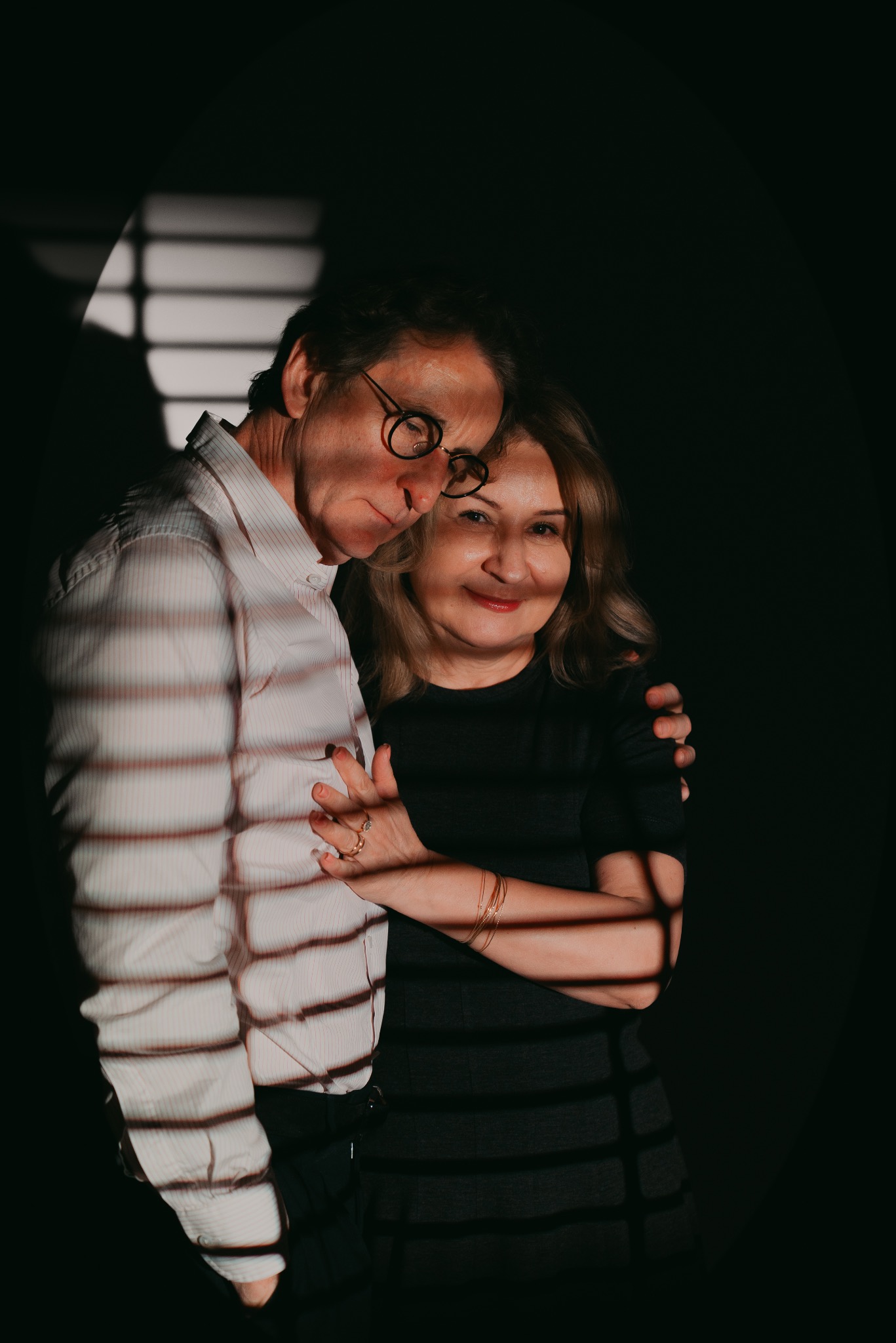 Portret pary w średnim wieku, mężczyzna w okularach i koszuli w paski obejmuje kobietę w czarnej sukience, na twarzach i ubraniach widoczne cienie od żaluzji.