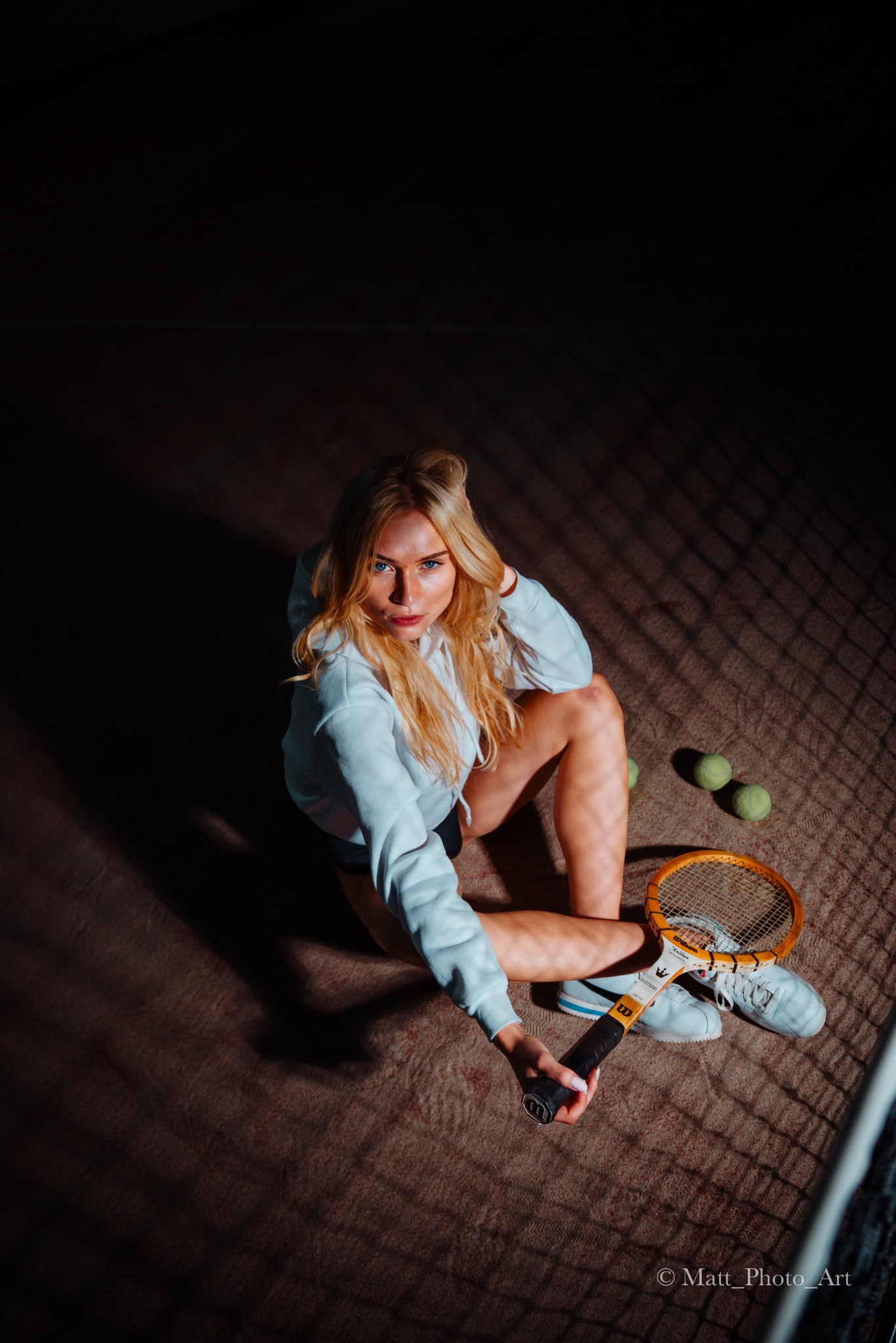 Kobieta w białej bluzie siedzi na korcie tenisowym, trzymając rakietę Wilson Knuckle, obok leżą trzy piłki tenisowe, oświetlenie z góry tworzy cienie kraty na podłożu.