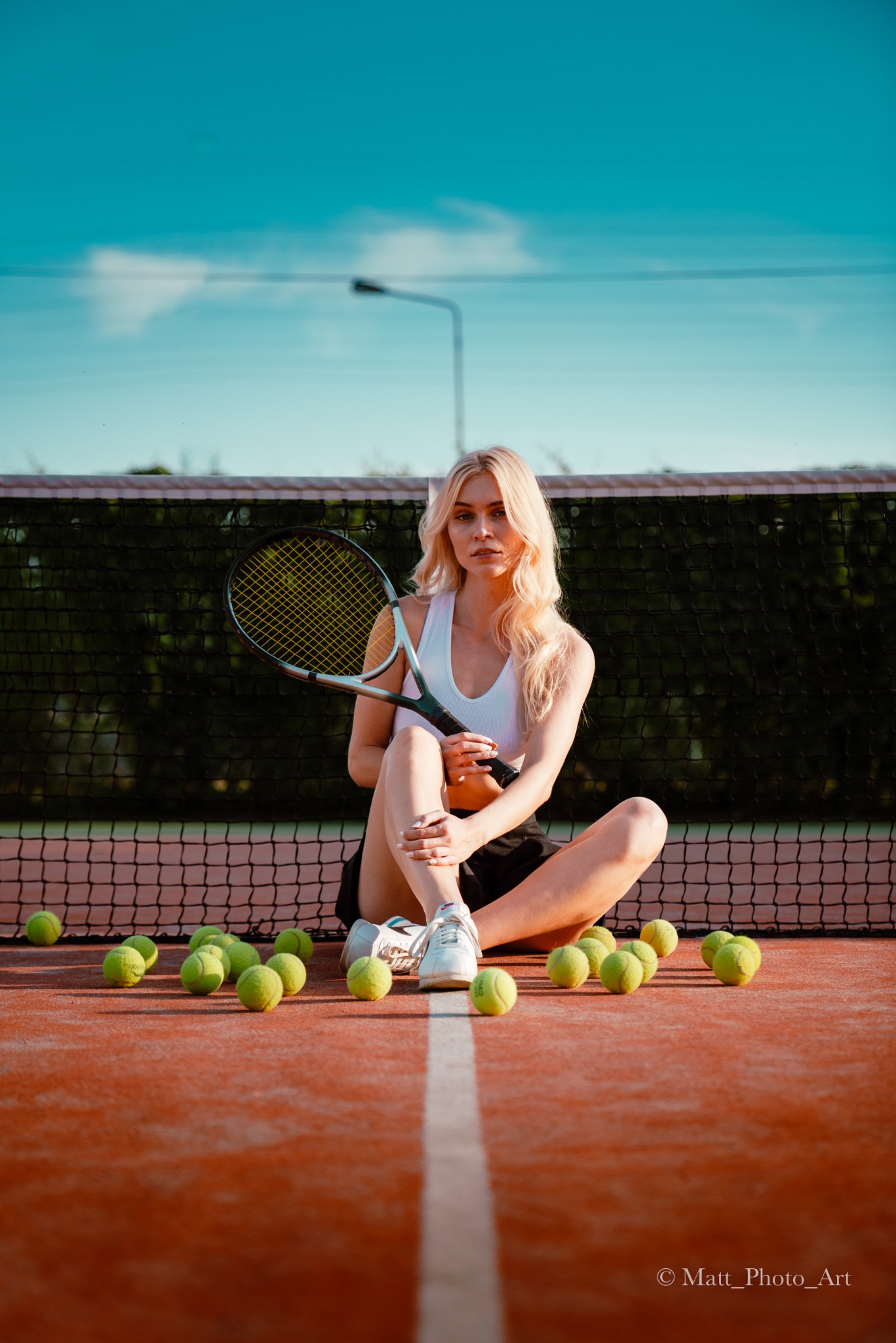 Blondwłosa kobieta siedzi na korcie tenisowym, trzyma rakietę, otoczona zielonymi piłkami, siatka w tle, oświetlenie popołudniowe.