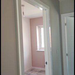 Smart Finishes - Świeżo pomalowane na różowo wnętrze pokoju widziane przez otwarte drzwi z białymi futrynami; podłoga zabezpieczona folią ochronną, surowa żarówka zwisa z sufitu.