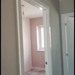 Świeżo pomalowane na różowo wnętrze pokoju widziane przez otwarte drzwi z białymi futrynami; podłoga zabezpieczona folią ochronną, surowa żarówka zwisa z sufitu.