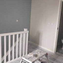 Smart Finishes - Wnętrze w trakcie odświeżania: ściana pomalowana częściowo na szaro, częściowo na biało, widoczna drabina malarska z pędzlem, biała balustrada schodów, podłoga zabezpieczona folią malarską.
