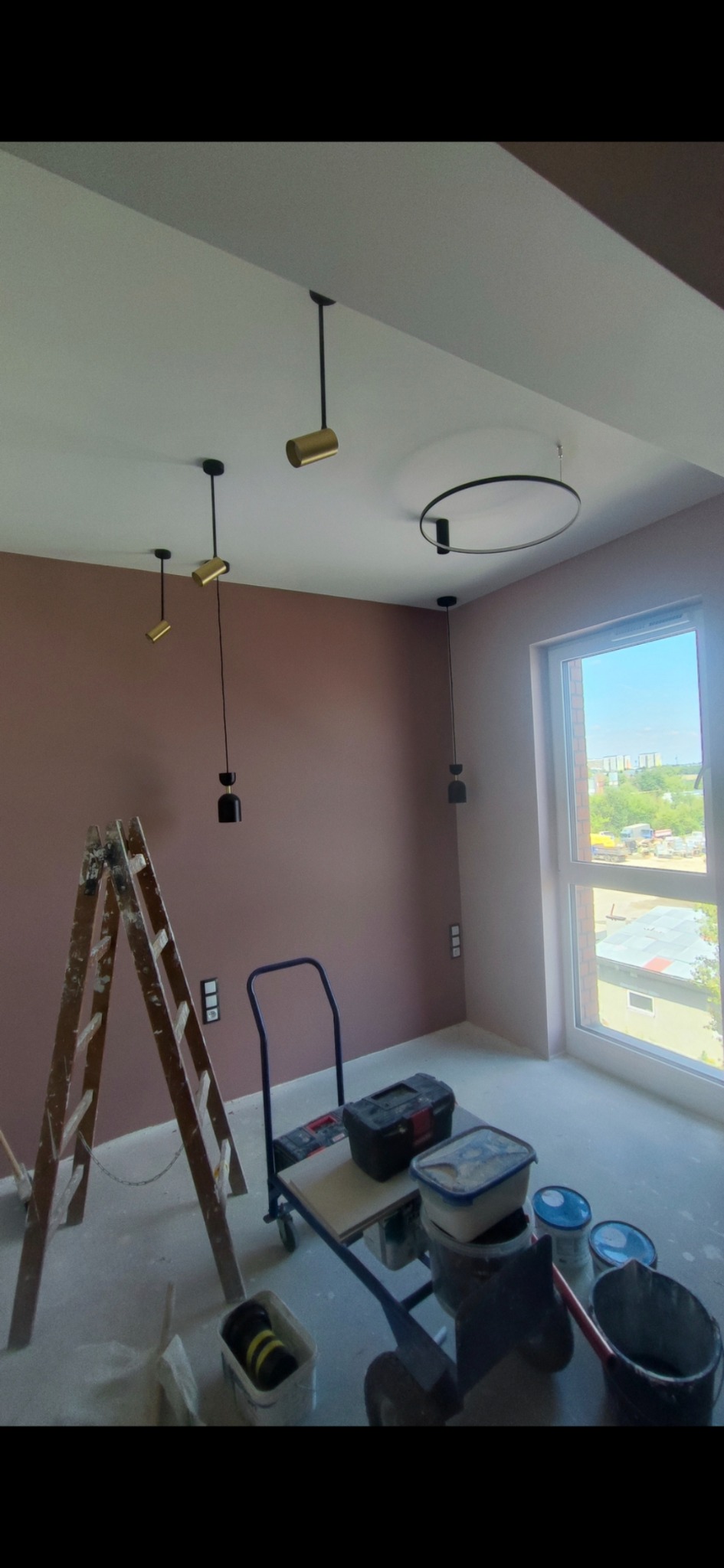 Pokój w trakcie malowania na kolor dusty rose, widoczna drabina, wózek z narzędziami, pędzle i wiaderka z farbą, nowoczesne oświetlenie sufitowe.