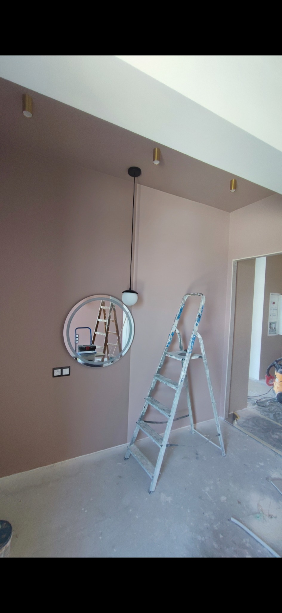 Pomieszczenie w trakcie malowania na dwa odcienie różu, widoczna drabina aluminiowa, okrągłe lustro z podświetleniem oraz lampa wisząca z kulistym kloszem.