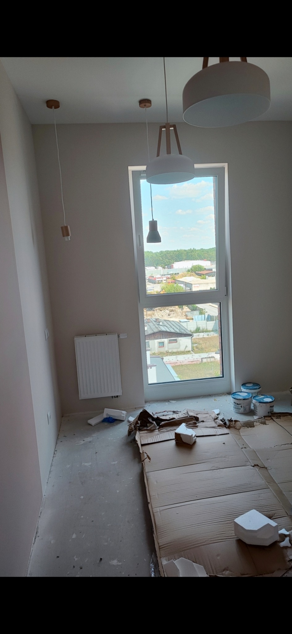 Pokój w trakcie prac wykończeniowych: świeżo pomalowane ściany w odcieniach beżu, zabezpieczona podłoga kartonem, widoczne puszki z farbą i nowoczesne lampy wiszące.