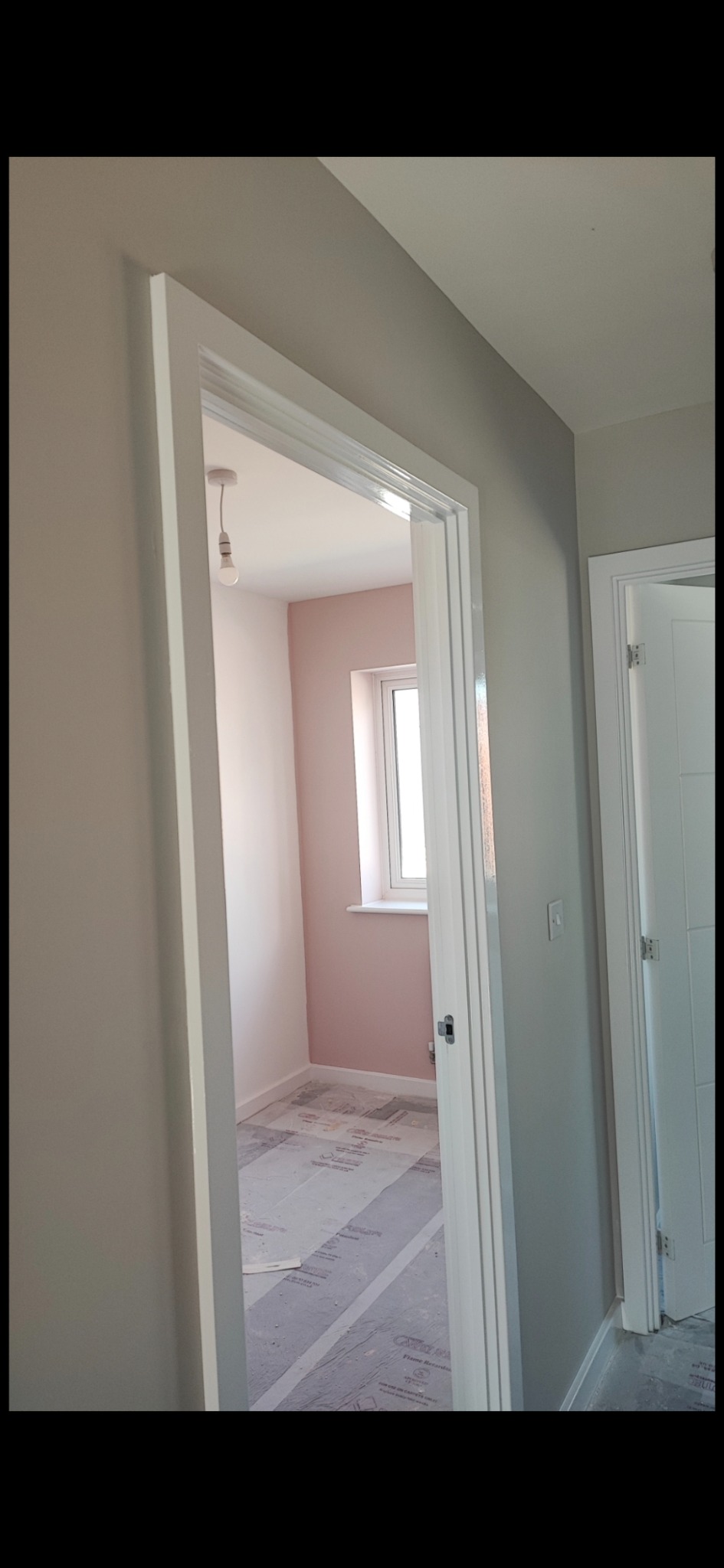 Świeżo pomalowane na różowo wnętrze pokoju widziane przez otwarte drzwi z białymi futrynami; podłoga zabezpieczona folią ochronną, surowa żarówka zwisa z sufitu.