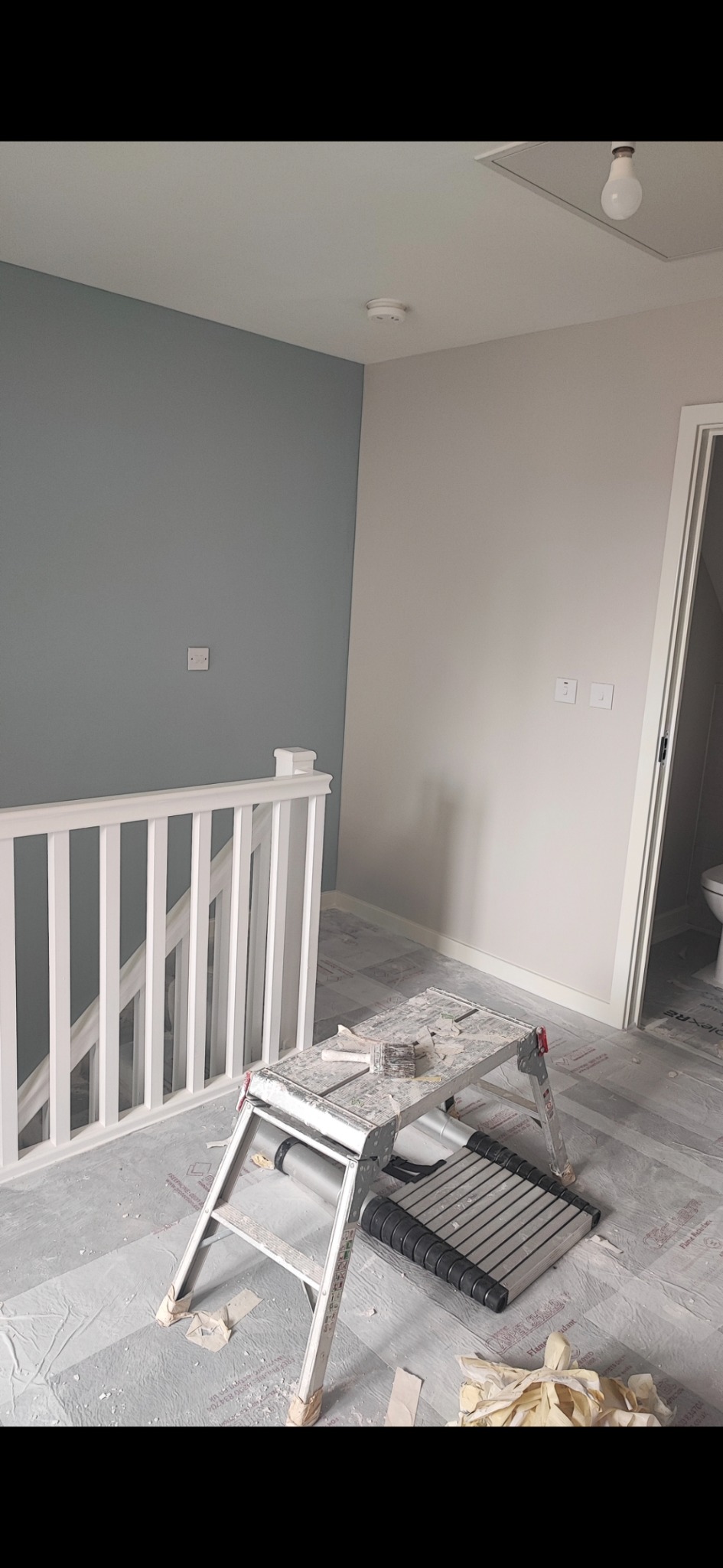 Wnętrze w trakcie odświeżania: ściana pomalowana częściowo na szaro, częściowo na biało, widoczna drabina malarska z pędzlem, biała balustrada schodów, podłoga zabezpieczona folią malarską.