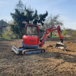 Czerwona mini koparka Wacker Neuson 28Z na wyrównanym, brązowym terenie. W tle domy i drzewa pod błękitnym niebem.