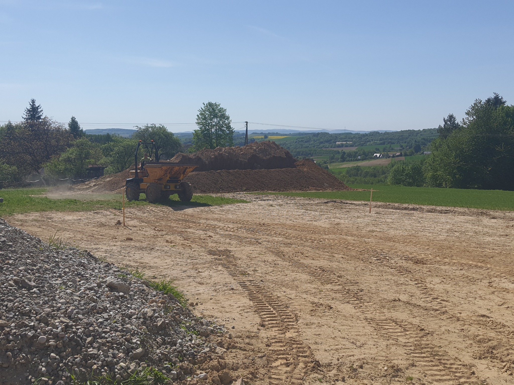 Żółta wywrotka w trakcie prac ziemnych na działce, z widocznymi śladami opon na piasku i stertą ziemi w tle, w słoneczny dzień.