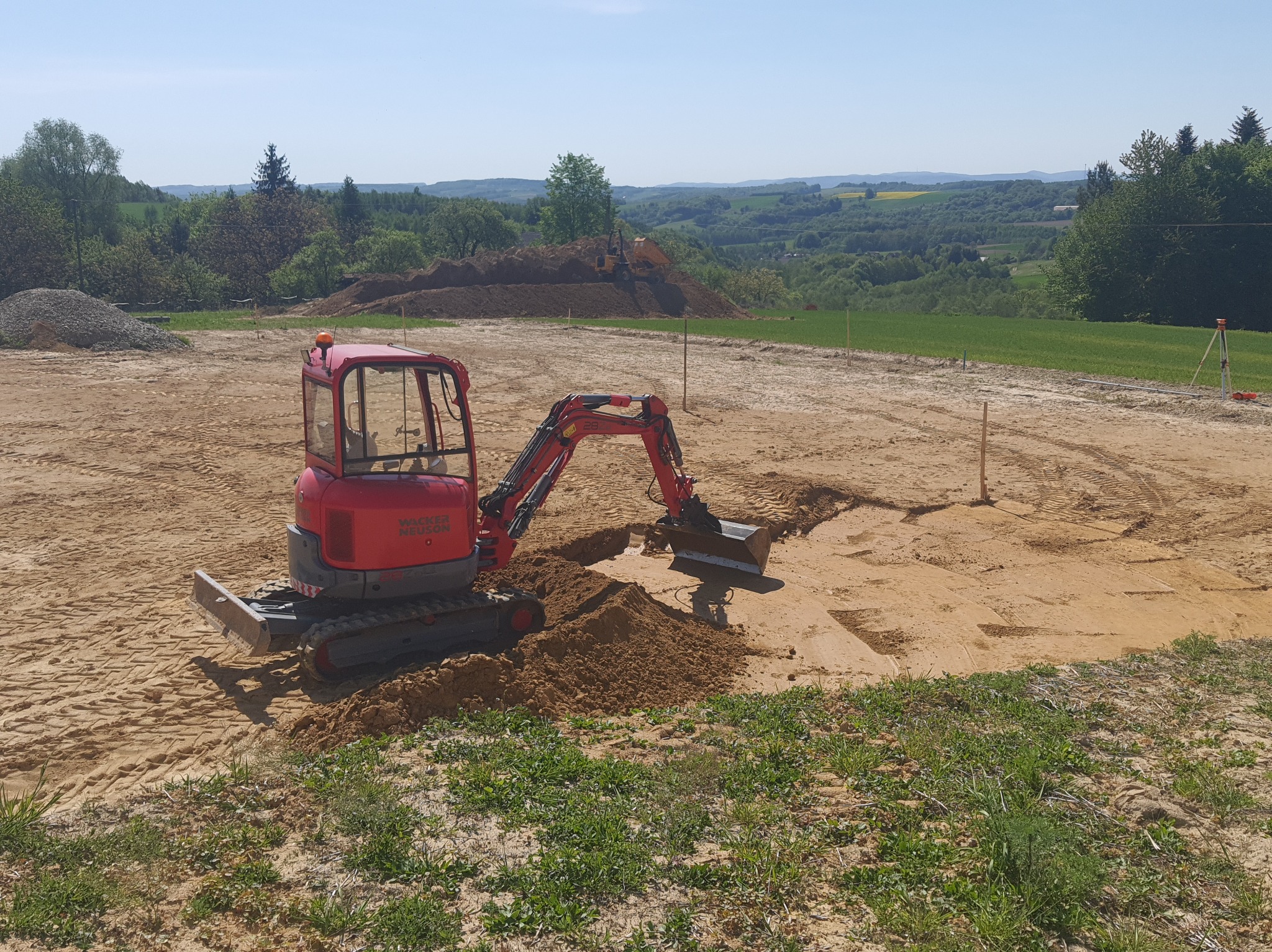 Czerwona mini koparka Wacker Neuson w trakcie wykopu rowu na placu budowy, widok na pagórkowaty krajobraz i stos materiałów budowlanych w tle.