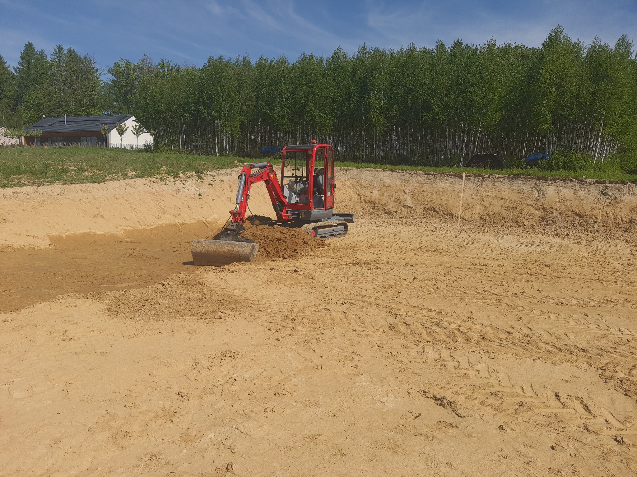 Czerwona mini koparka w trakcie prac ziemnych na piaszczystym terenie, prawdopodobnie wykopywanie stawu. W tle drzewa i budynek.