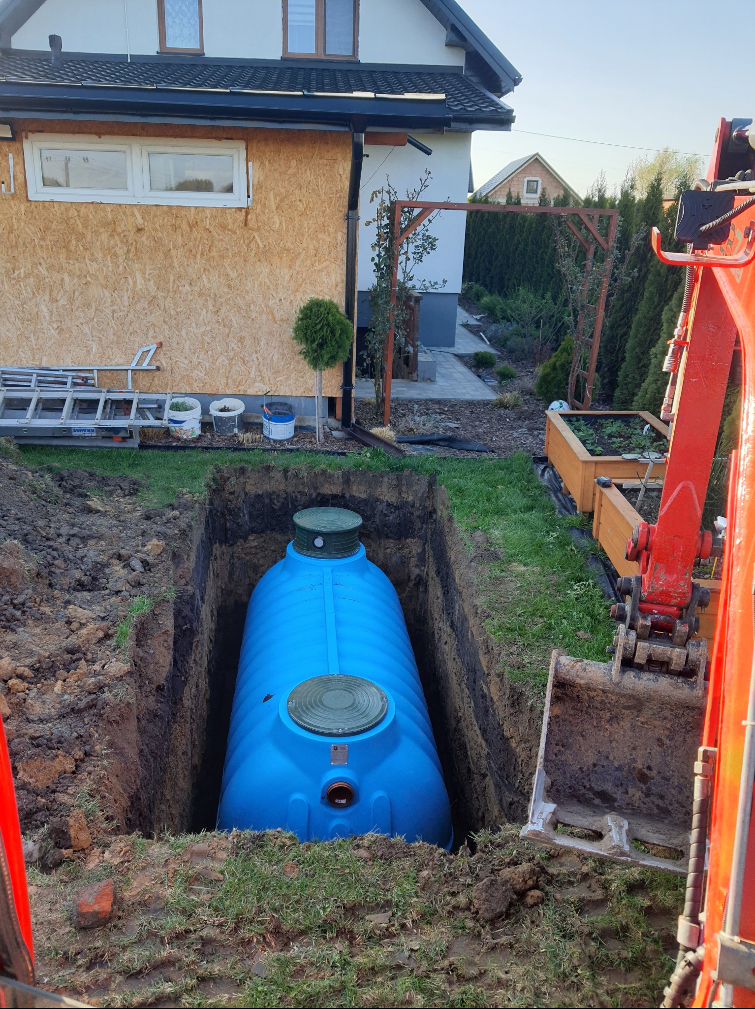 Niebieski zbiornik na deszczówkę w wykopie, widoczny fragment koparki, trawa, dom w tle. Montaż podziemnego zbiornika na wodę deszczową w ogrodzie.