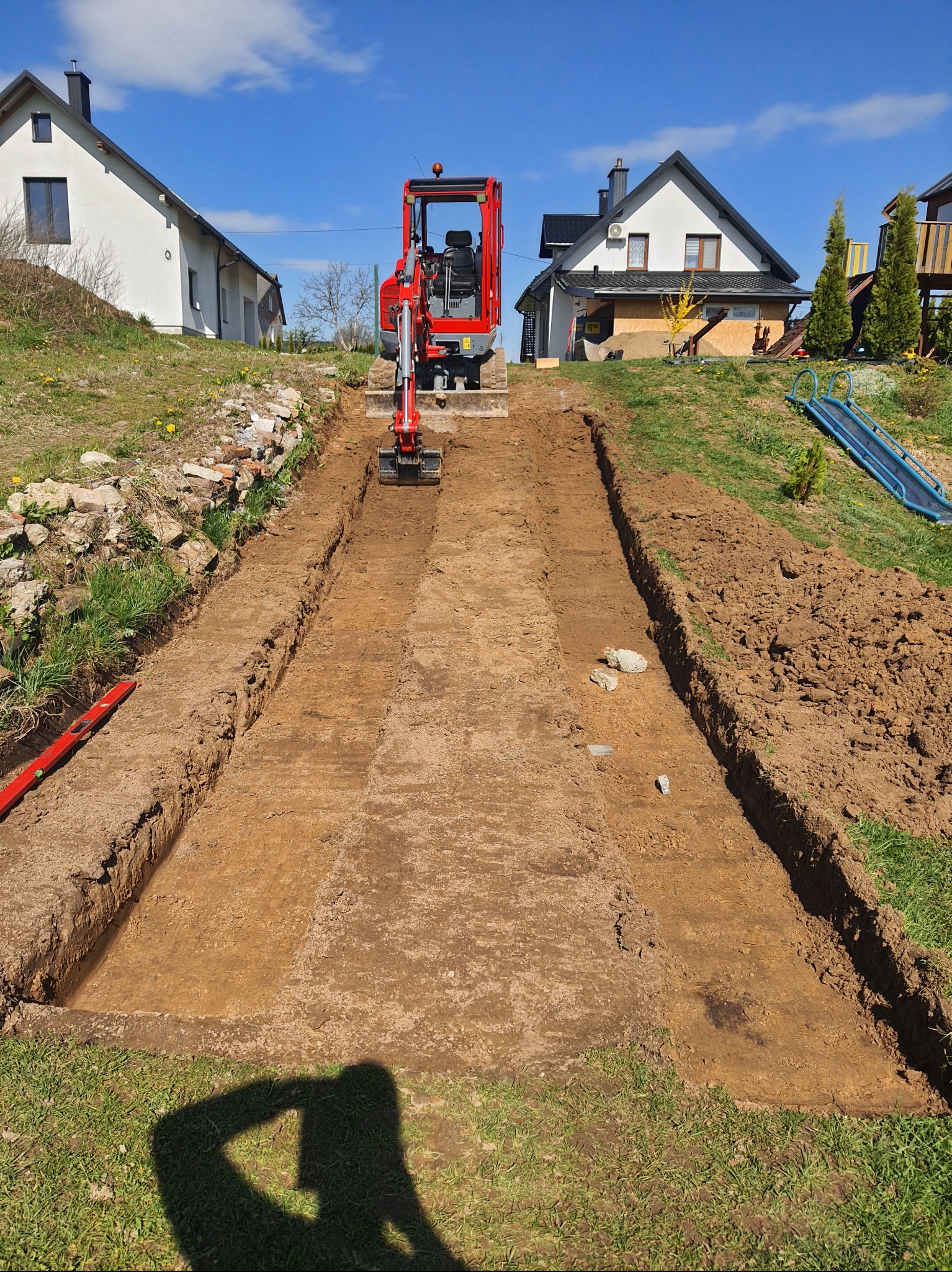 Czerwona mini koparka na działce, przygotowuje wykop pod fundamenty. Widoczne wykopane rowy, poziomica i budynki w tle. Błękitne niebo.
