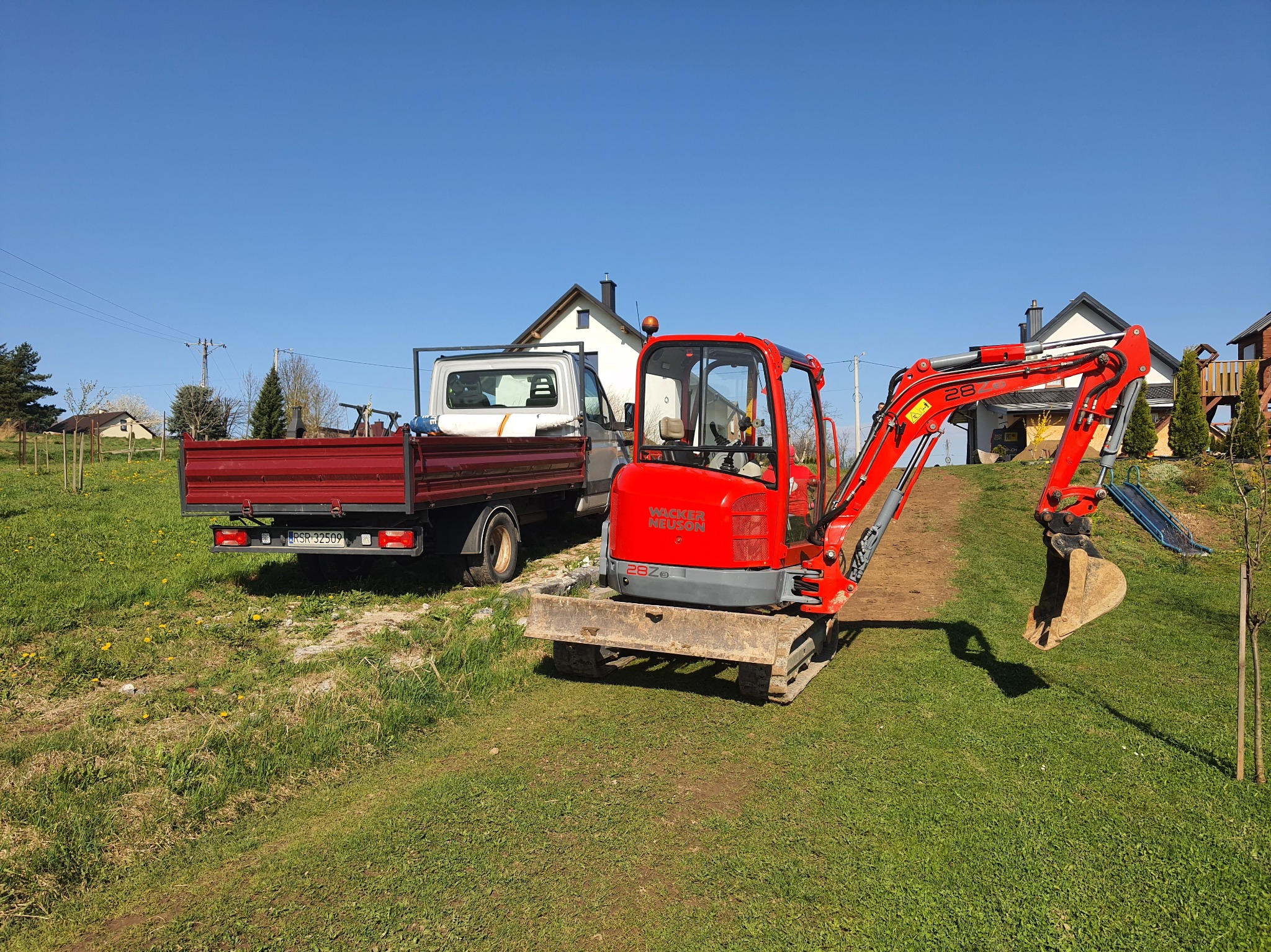 Czerwona mini koparka Wacker Neuson 28Ze na trawie obok wywrotki z czerwoną skrzynią, w tle domy jednorodzinne i błękitne niebo.